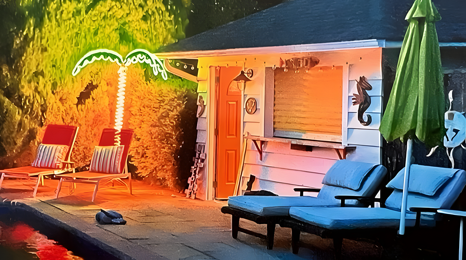 View of a backyard scene at dusk with a clam shell shaped neon sign, a palm tree, two lounge chairs with striped cushions, a sunbed, a house with a window and door, and an outdoor umbrella