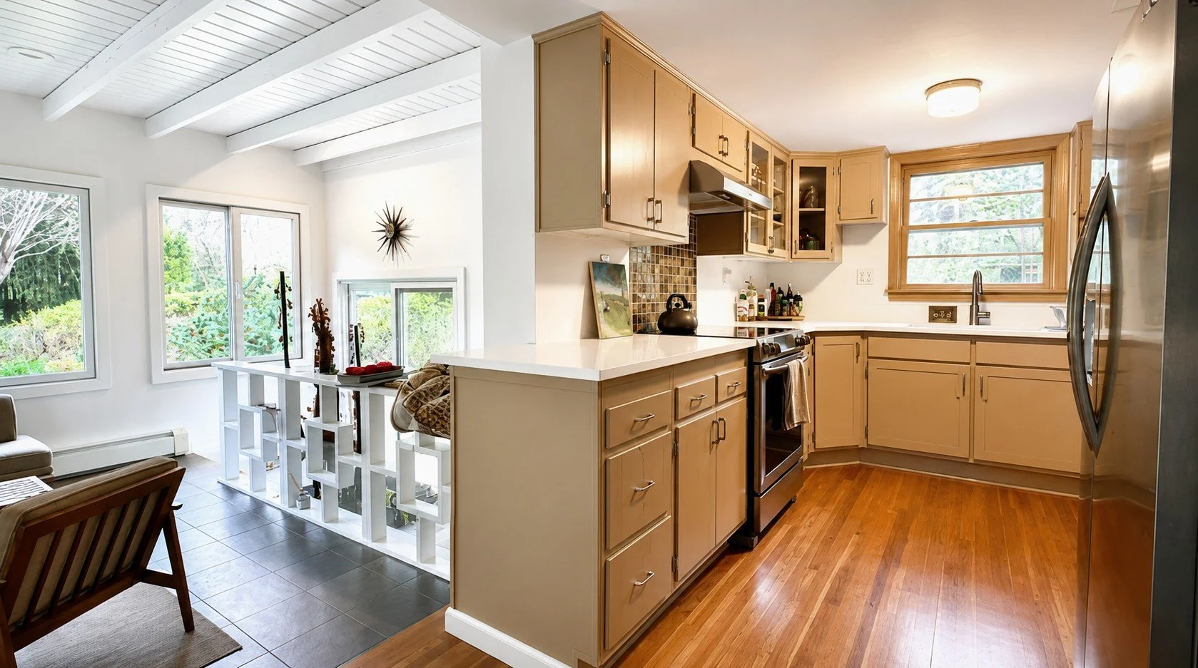The image shows a kitchen with beige cabinets, a window above the sink, and a white countertop. Adjacent to the kitchen, there is a small sitting area with large windows, a lamppost inside, and some décor on a white partition. The flooring transition