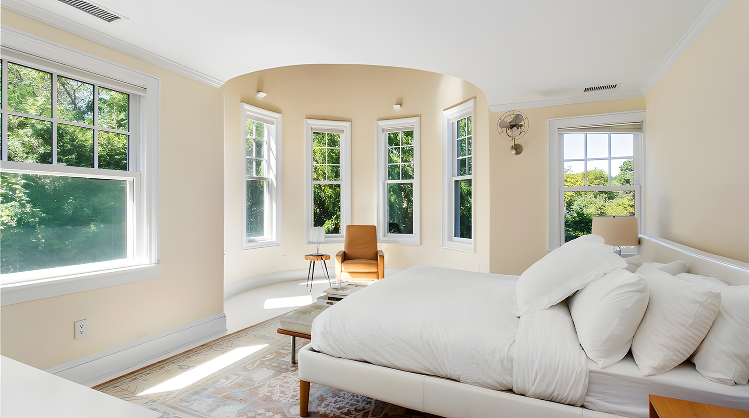 Bright bedroom with five large windows, a beige armchair, a small wooden side table, a bed with white linens, a table lamp, and a wall fan, with greenery visible outside.