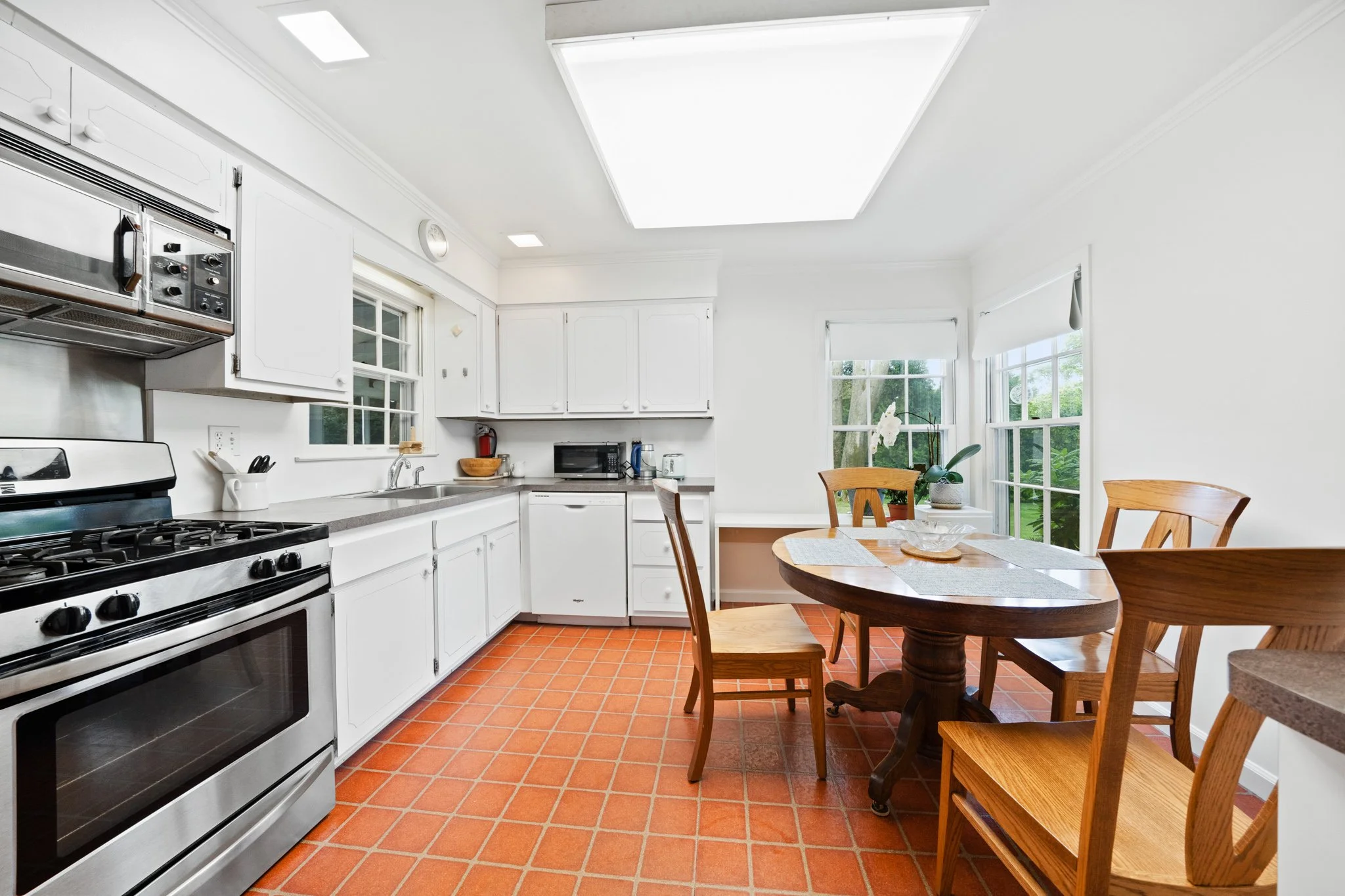 Bright kitchen with white cabinets, stainless steel stove, black microwave, and wooden dining table with chairs, large windows, and a skylight overhead.