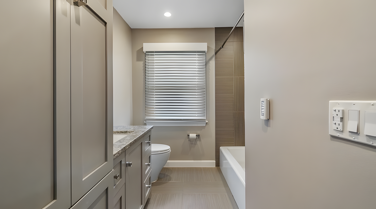 Modern bathroom with gray cabinetry, a granite countertop, a toilet, a window with blinds, a bathtub, and beige tile flooring.