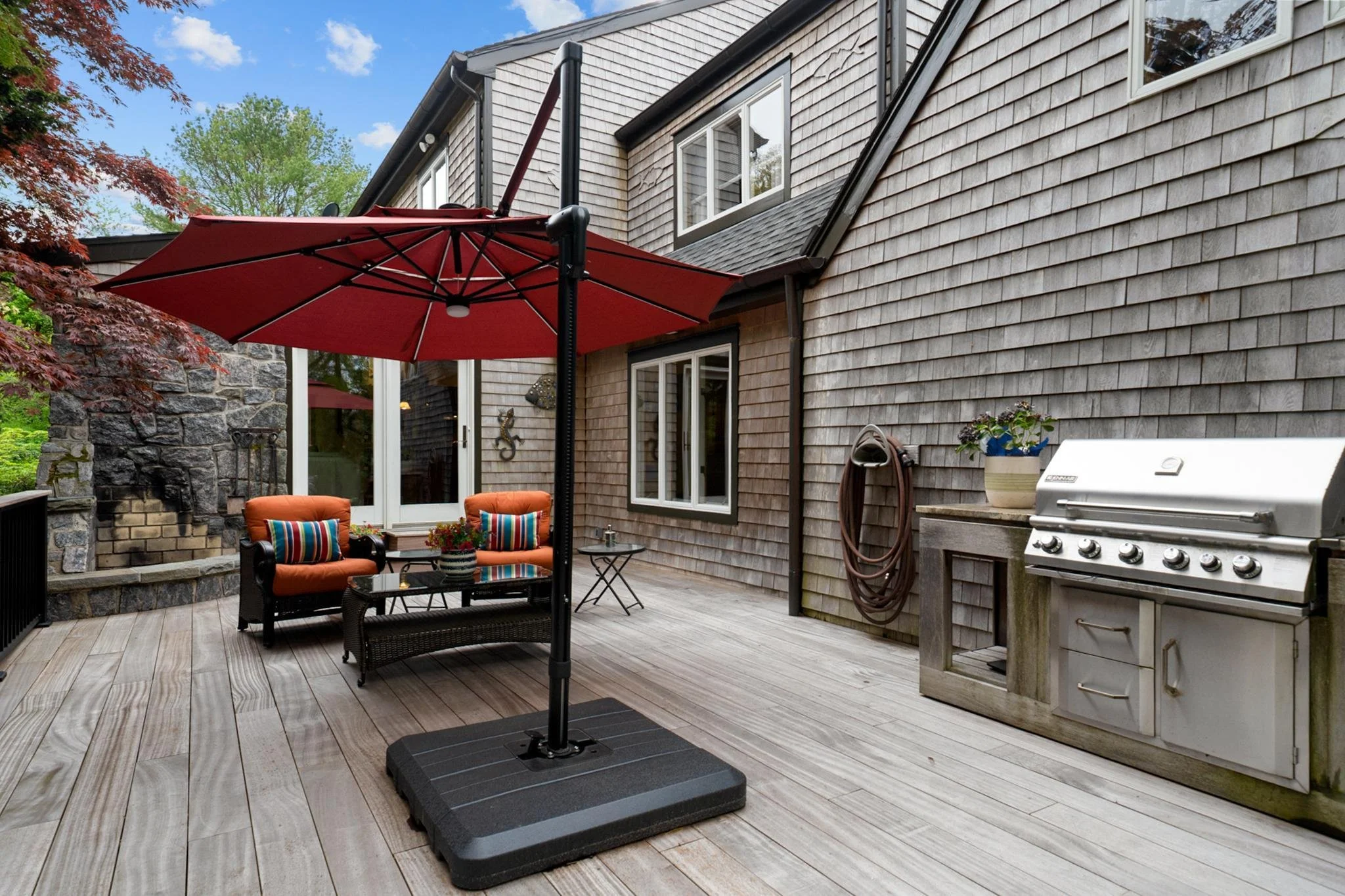 A backyard patio with wooden flooring, a red patio umbrella, black wicker furniture with orange cushions, a small side table with flowers, a gas grill, and house siding with four windows and a downspout, surrounded by trees and cloudy sky.