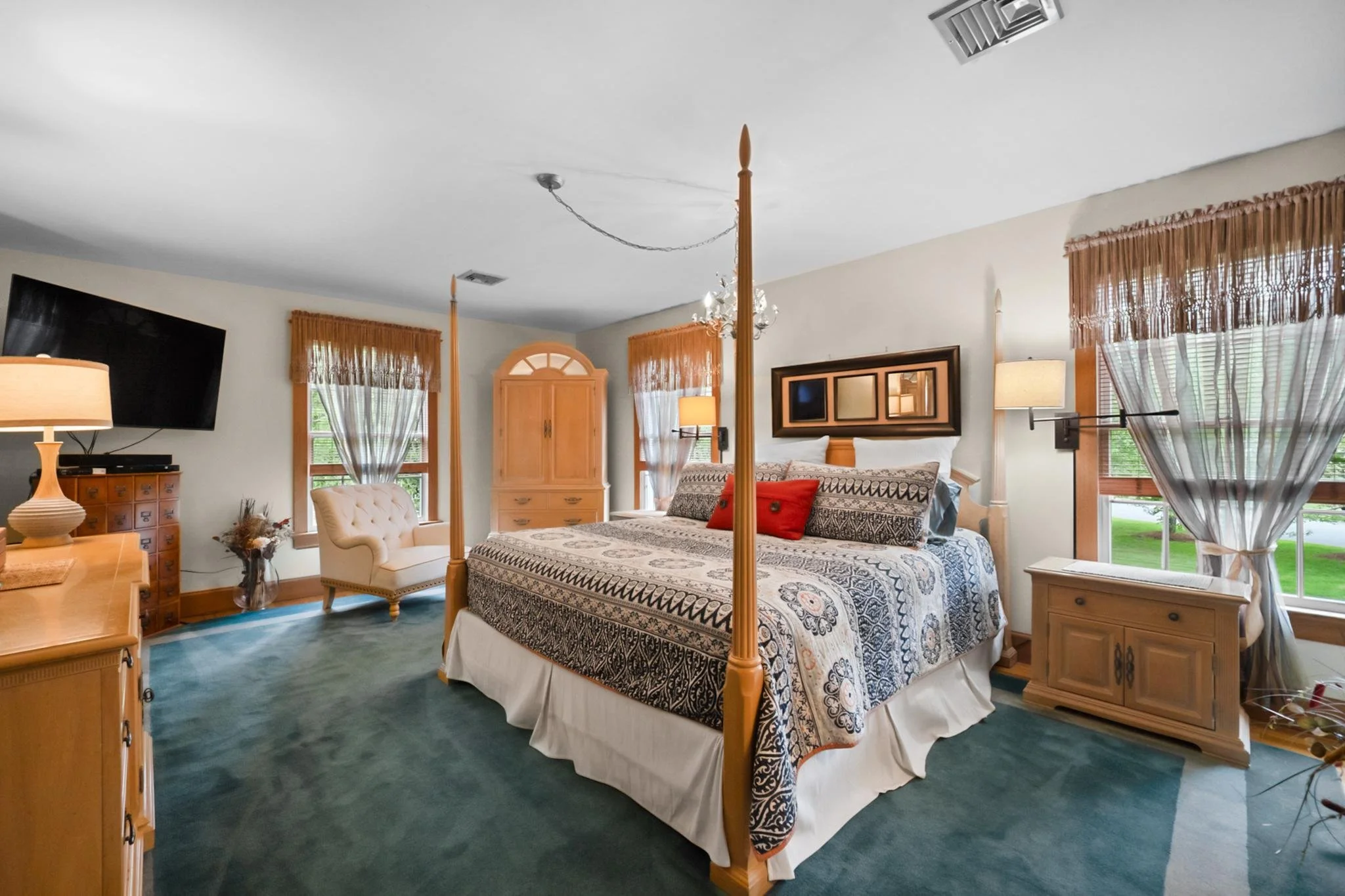 A bedroom with a four-poster bed, a chest of drawers, a TV on a stand, a beige armchair, a nightstand, and large windows with wooden blinds and sheer curtains, illuminated by natural light and two table lamps.