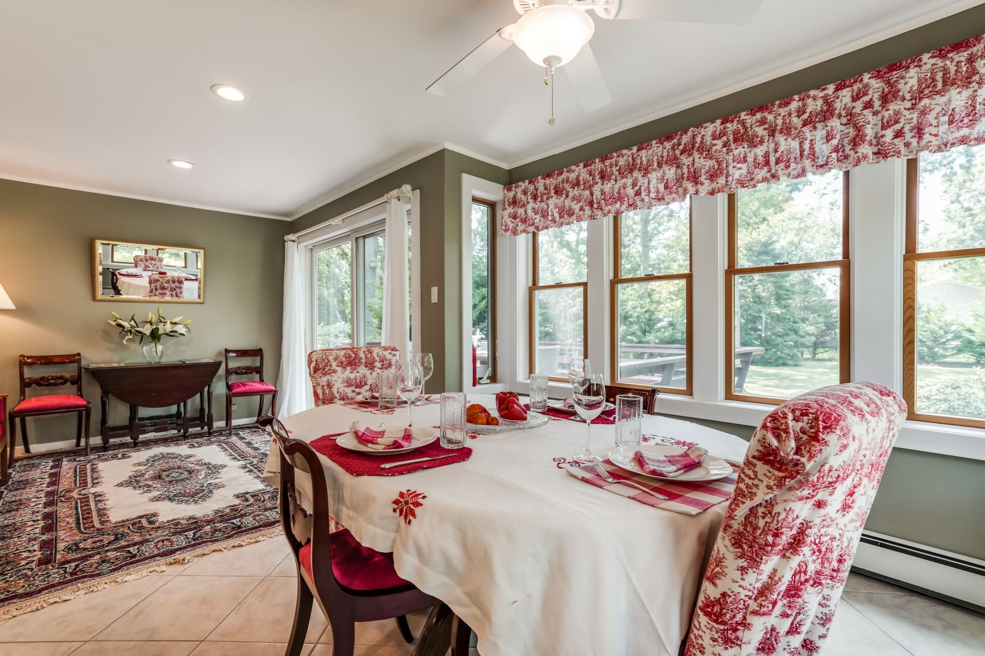 A dining room with a table set for a meal, surrounded by chairs with red and white patterned upholstery. Large windows with a red and white patterned valance and white curtains let in sunlight, showing a view of trees outside. A ceiling fan with ligh