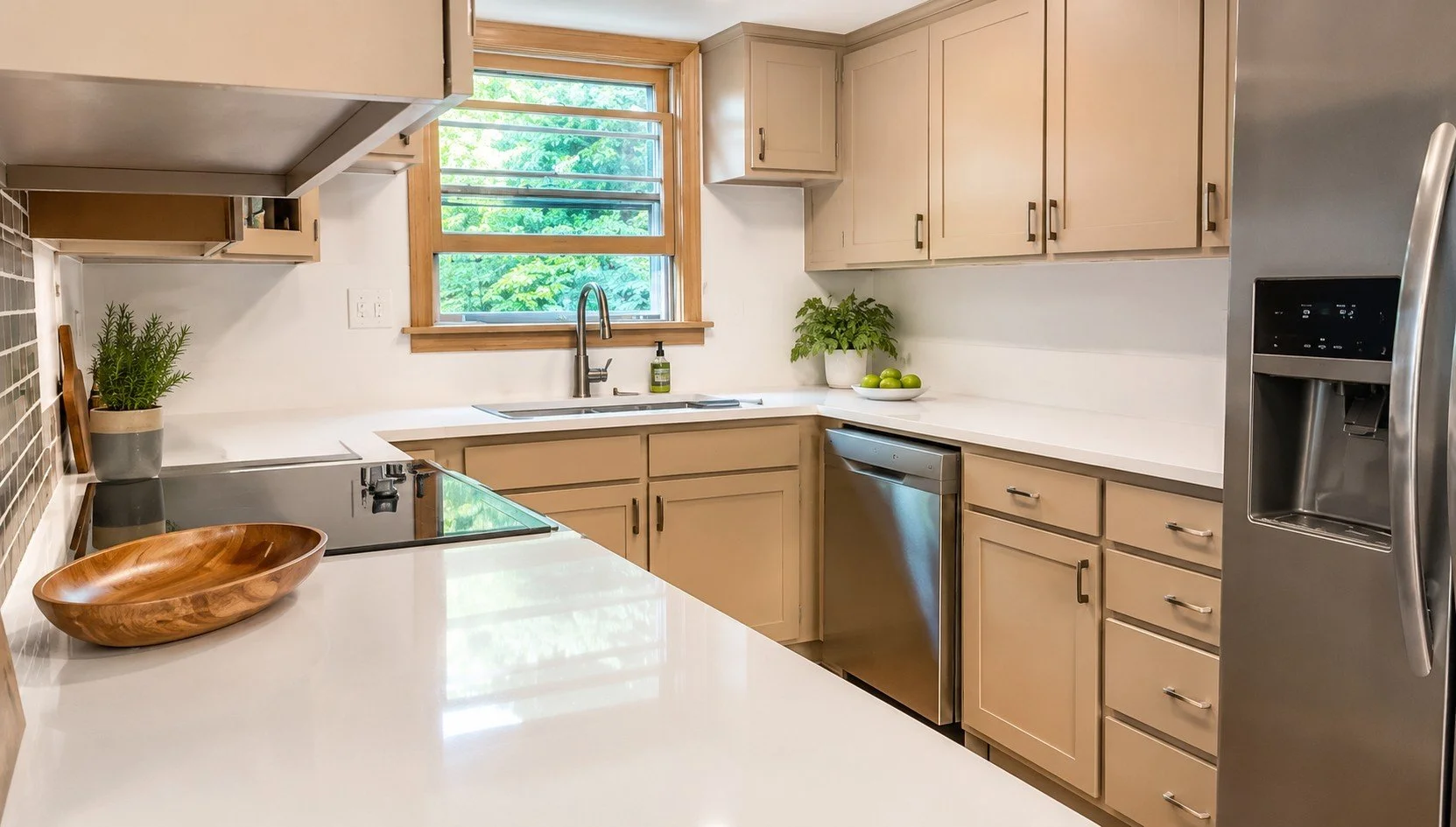 Modern kitchen with beige cabinets, white countertops, a stainless steel refrigerator, dishwasher, and sink, with a window showing greenery outside, and decorative items like potted plants and a wooden bowl.