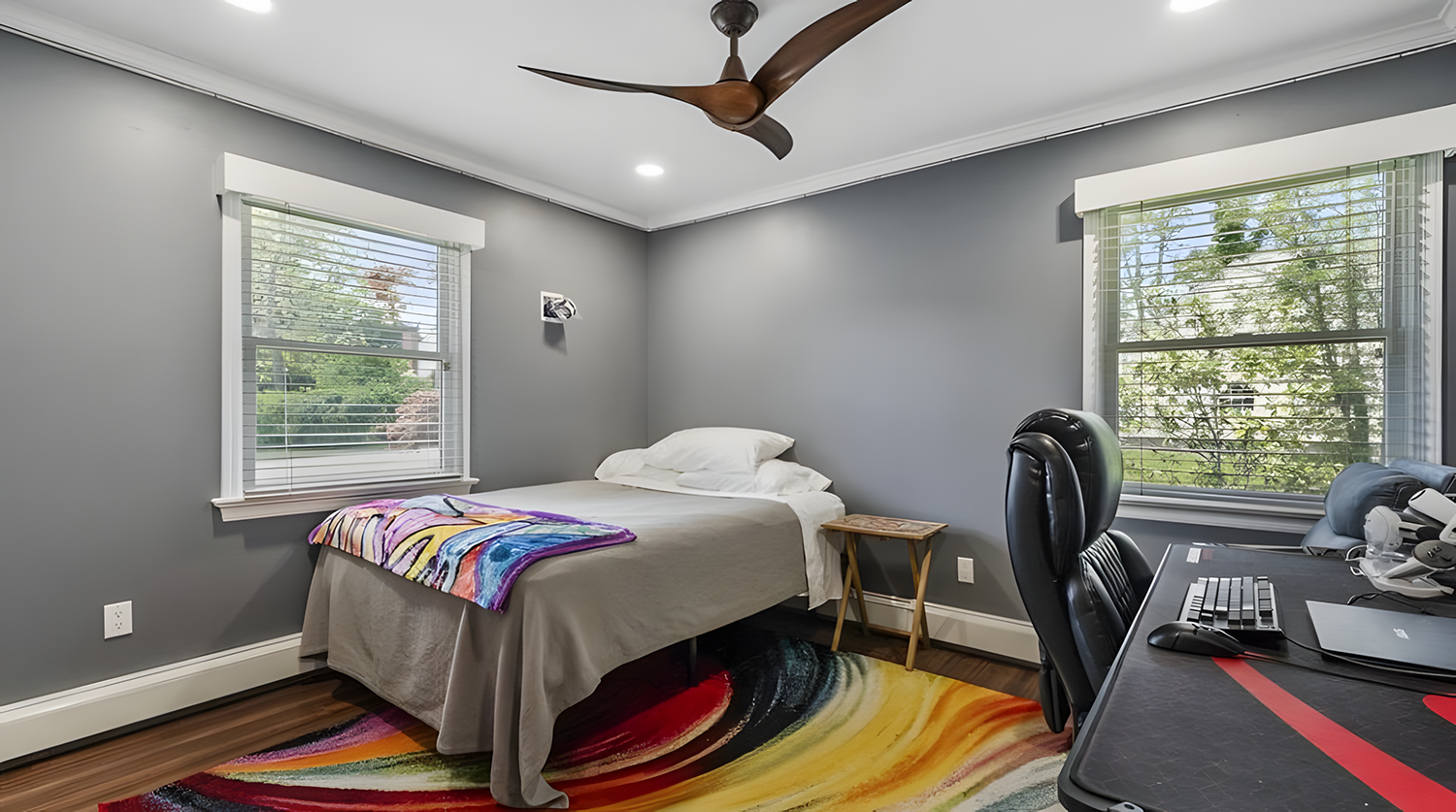 A bedroom with gray walls, a bed with white pillows and blanket, two windows with blinds, a small side table, a colorful abstract rug, and a desk with a computer, keyboard, mouse, and headphones.