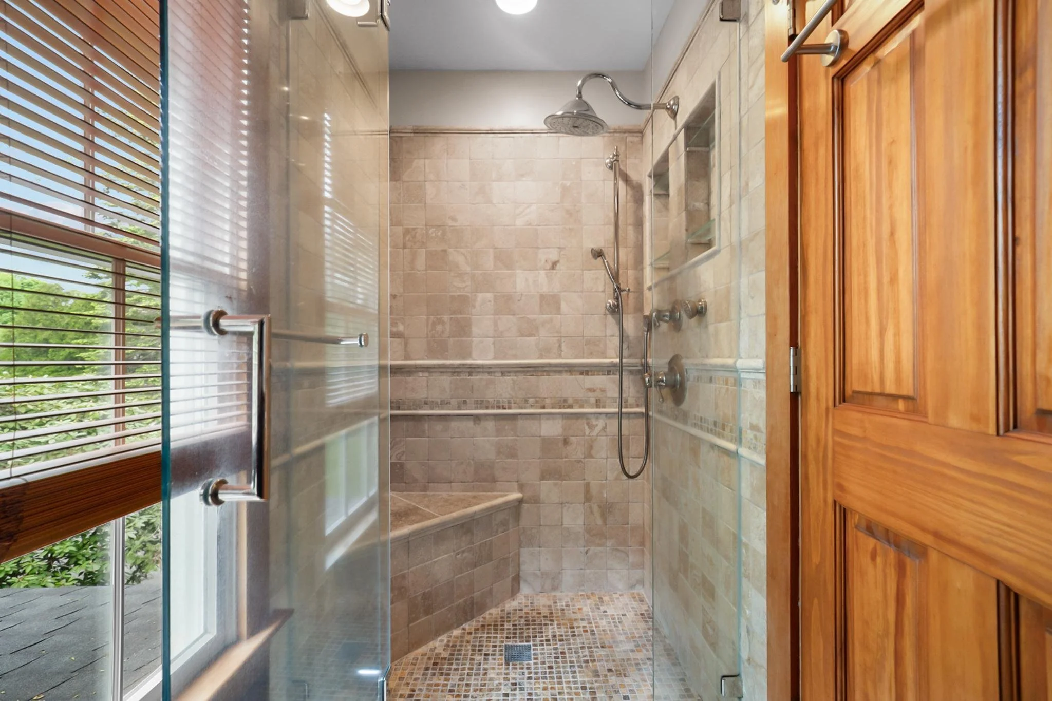 A bathroom shower area with beige tiled walls and a glass door. There is a large rain shower head and a smaller handheld shower head with controls below. A small built-in shower niche is visible. Wooden window blinds are on the left, and a wooden cab