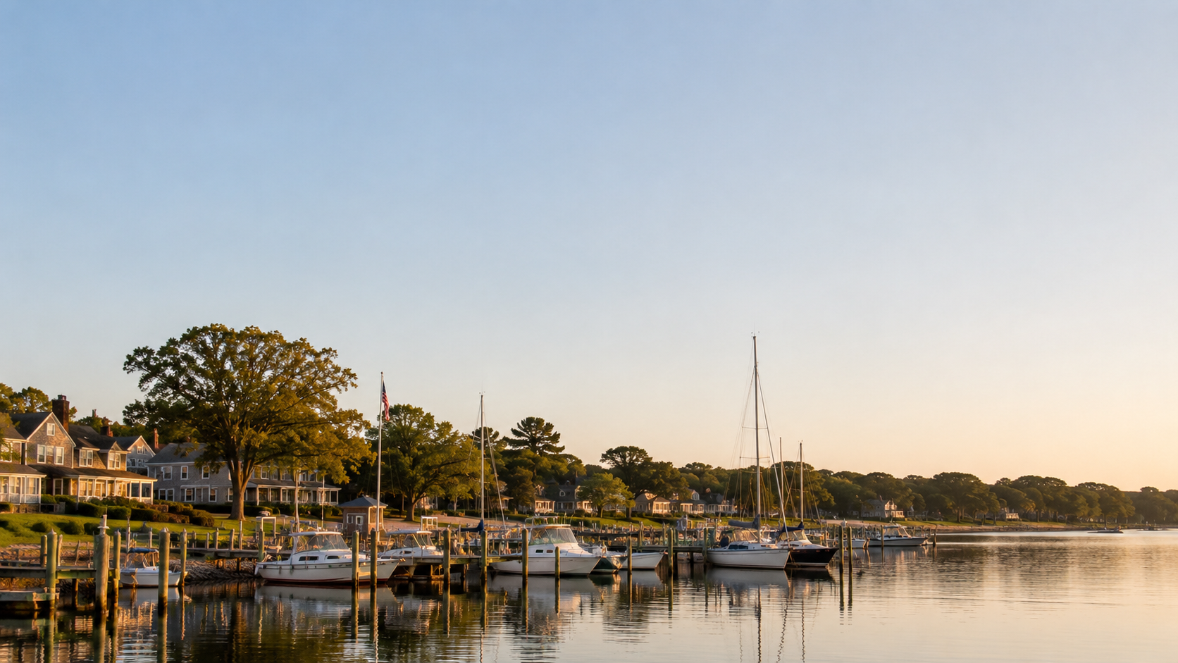A calm waterfront with several sailboats docked along a wooden pier, houses and lush trees in the background at sunset.