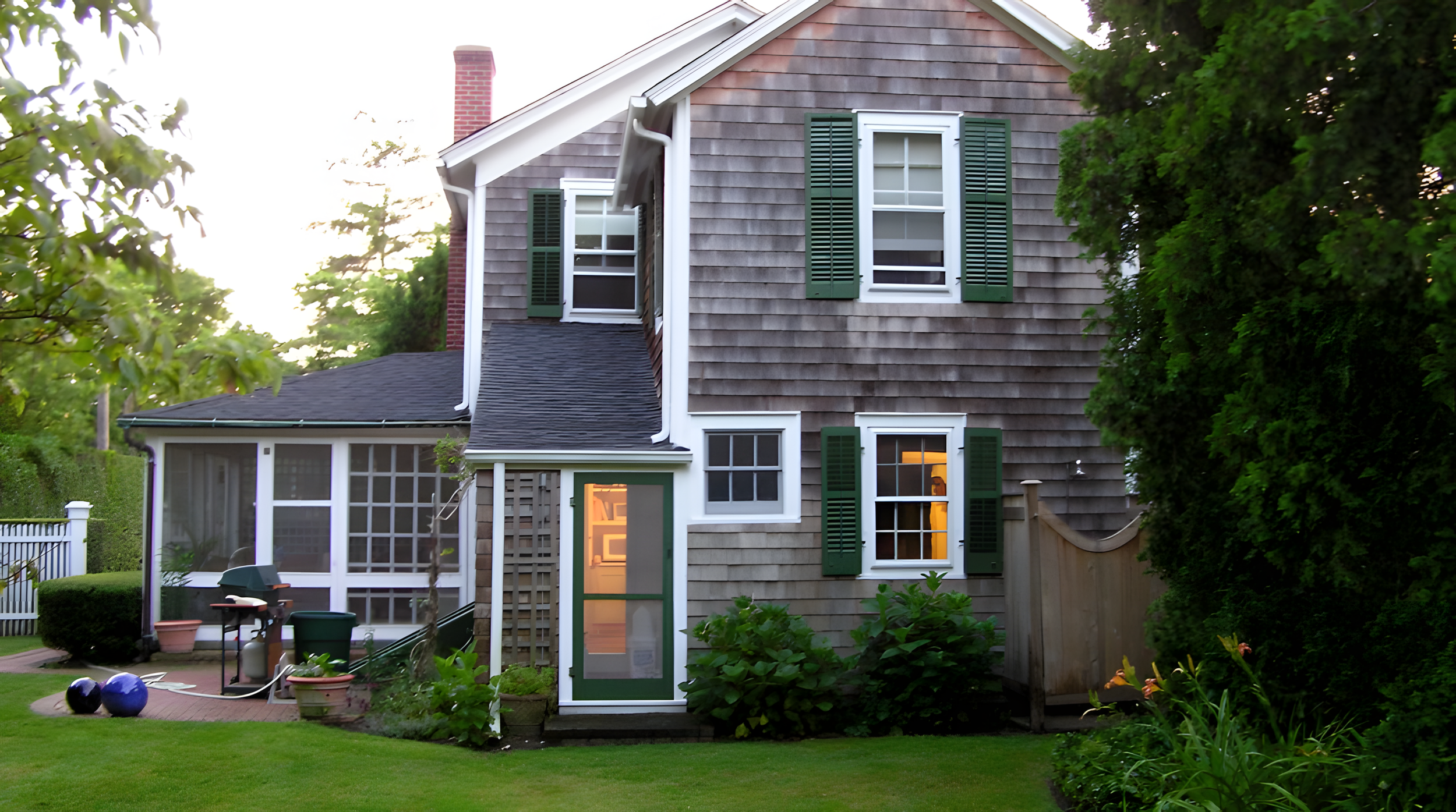 A two-story house with gray shingle siding, multiple windows with green shutters, and a chimney, surrounded by green trees and a well-maintained lawn.