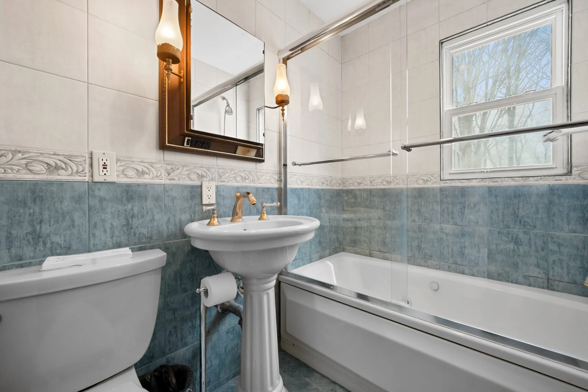 A bathroom with white and blue tiles, a toilet, a pedestal sink with a gold faucet, a mirror, and a bathtub with a sliding glass shower door. There is a window showing trees outside.
