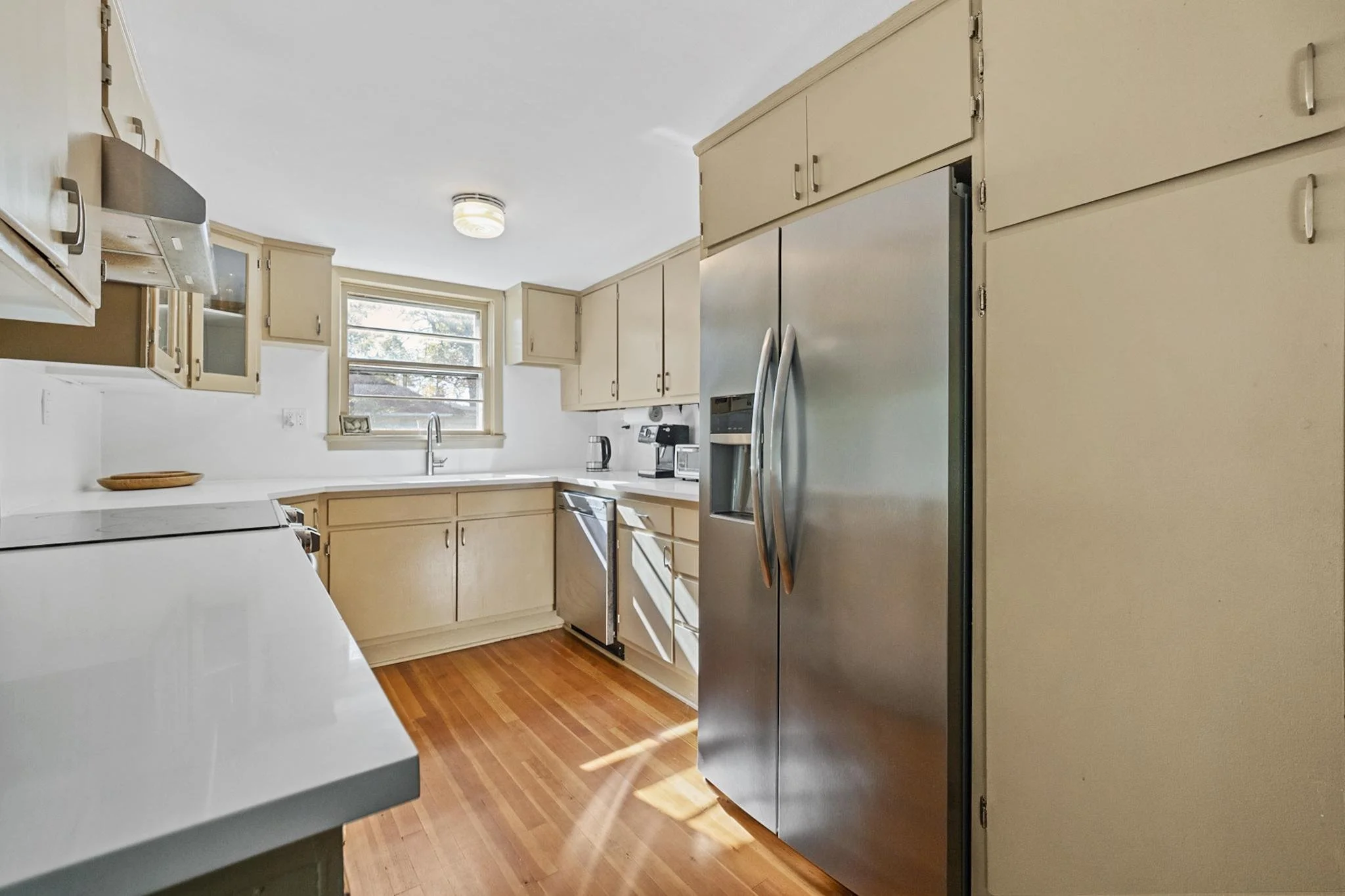 Kitchen with beige cabinets, stainless steel refrigerator, white countertop, wooden floor, window above sink, microwave, and coffee maker.