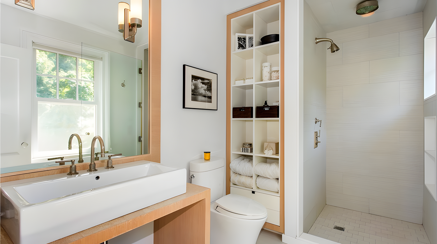 Modern bathroom with a large mirror, white sink, wooden accents, a window, a toilet, and a shower area with a wall-mounted showerhead.