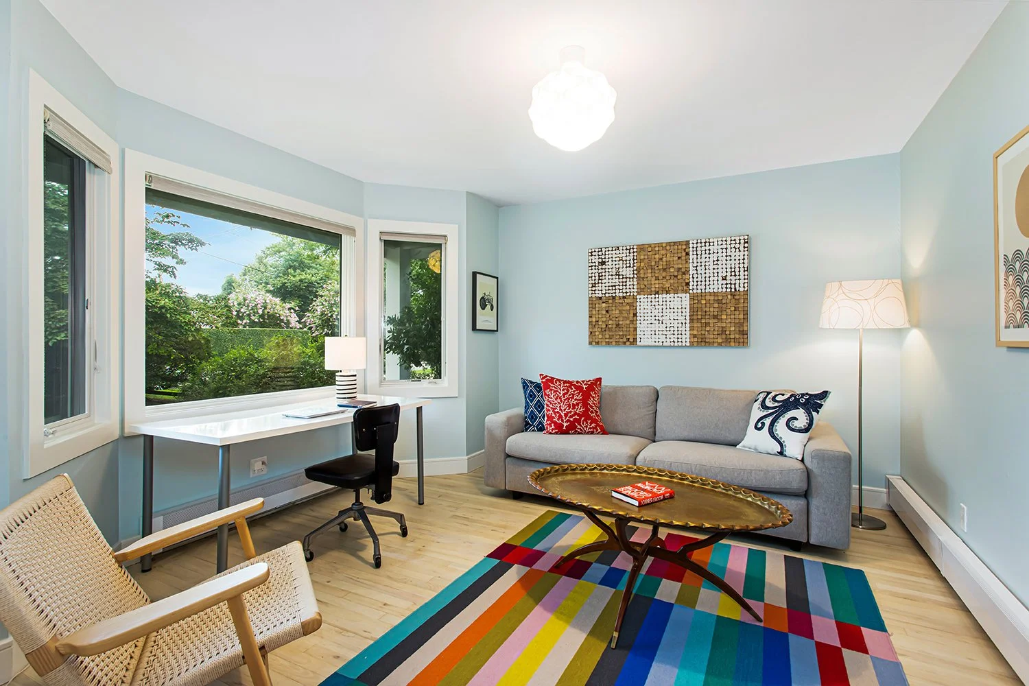 Living room with light blue walls, a grey sofa with colorful pillows, a wooden coffee table, a multicolored striped rug, a floor lamp, and a desk by the window with a chair, large windows showing greenery outside.