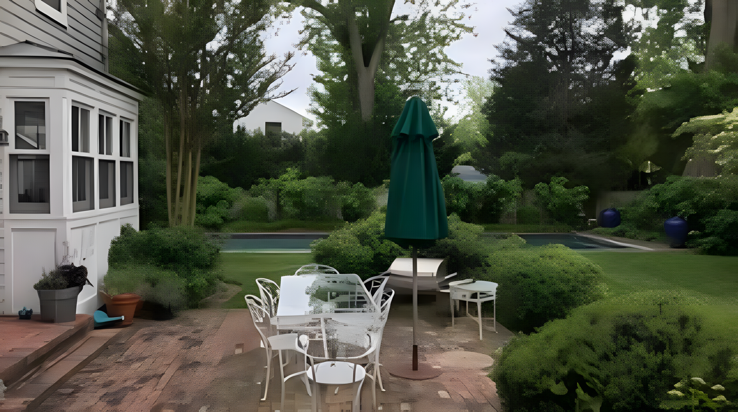 A backyard patio with white outdoor furniture, a green umbrella, lush green trees, and a swimming pool surrounded by bushes. A white house with large windows is visible on the left.