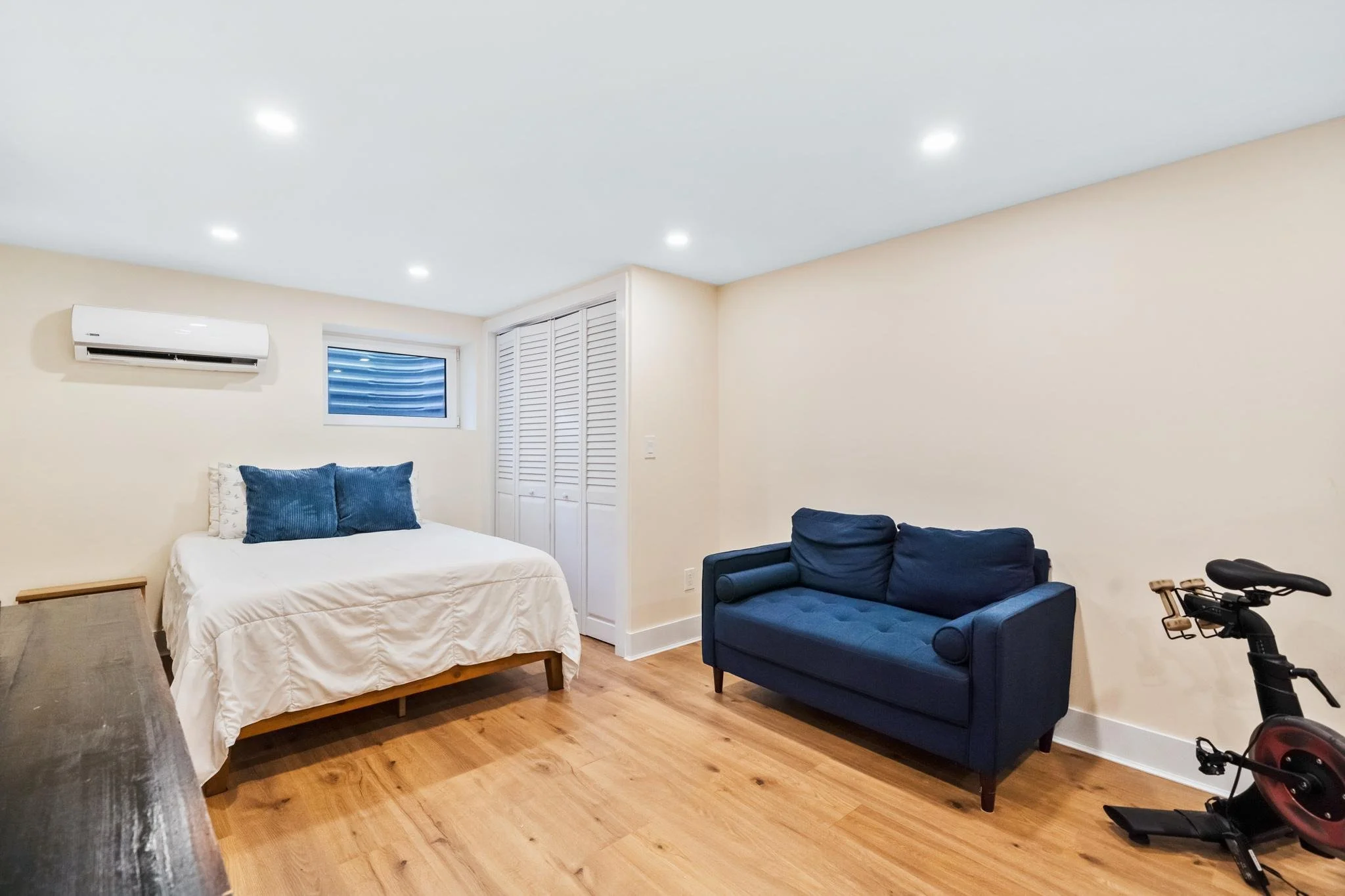 A bedroom with a white bed, blue cushions, a window, a closet with louvered doors, a navy blue sofa, and an exercise bike on a hardwood floor.