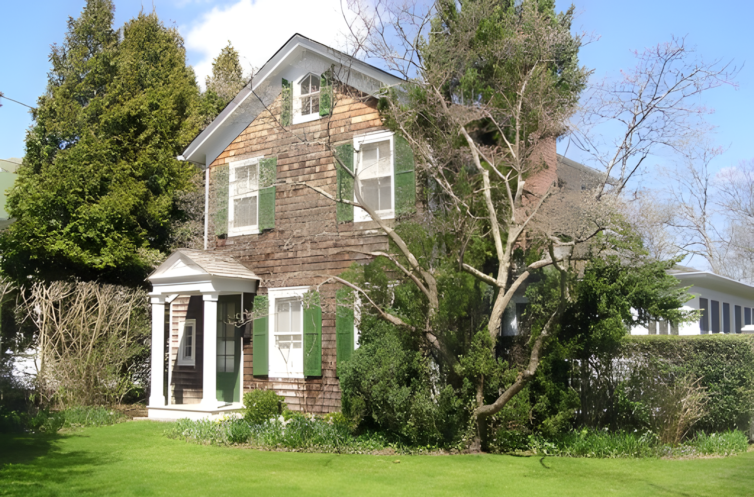 A multi-story house with weathered brown shingles, white trim, and green shutters, surrounded by a green lawn, shrubs, and large trees under a blue sky.