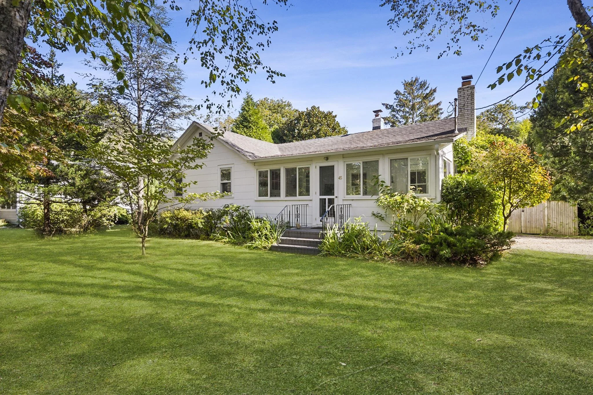 A white house with a screened porch, surrounded by a lush green lawn, trees, and bushes, with a clear blue sky overhead.