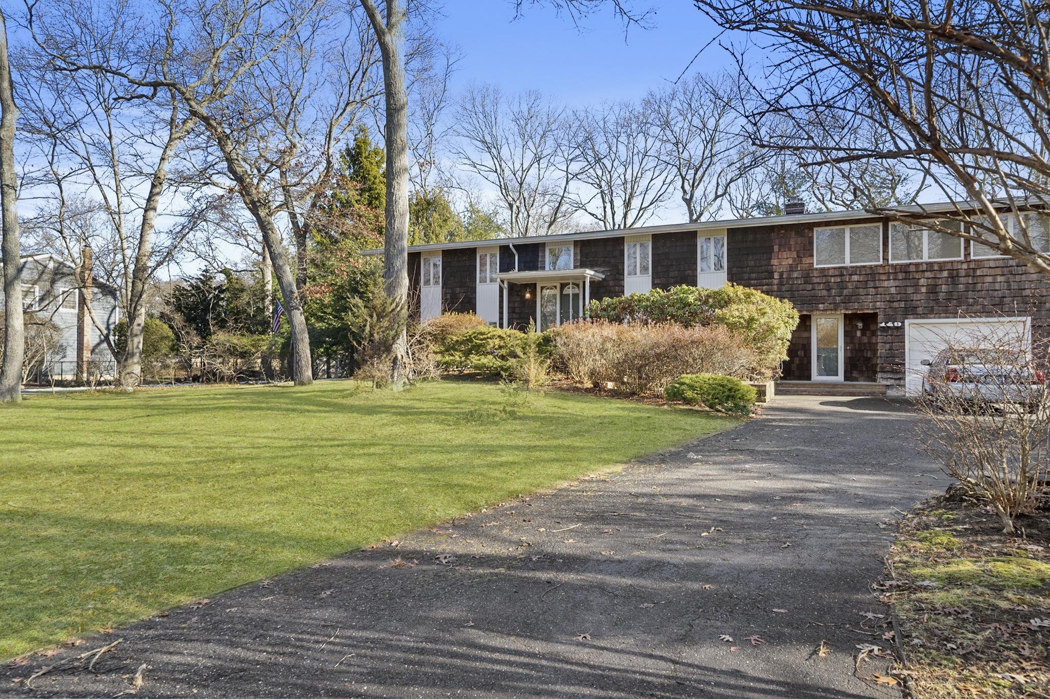 A suburban house with brown shingle siding, surrounded by leafless trees and a well-maintained lawn, under a clear blue sky.