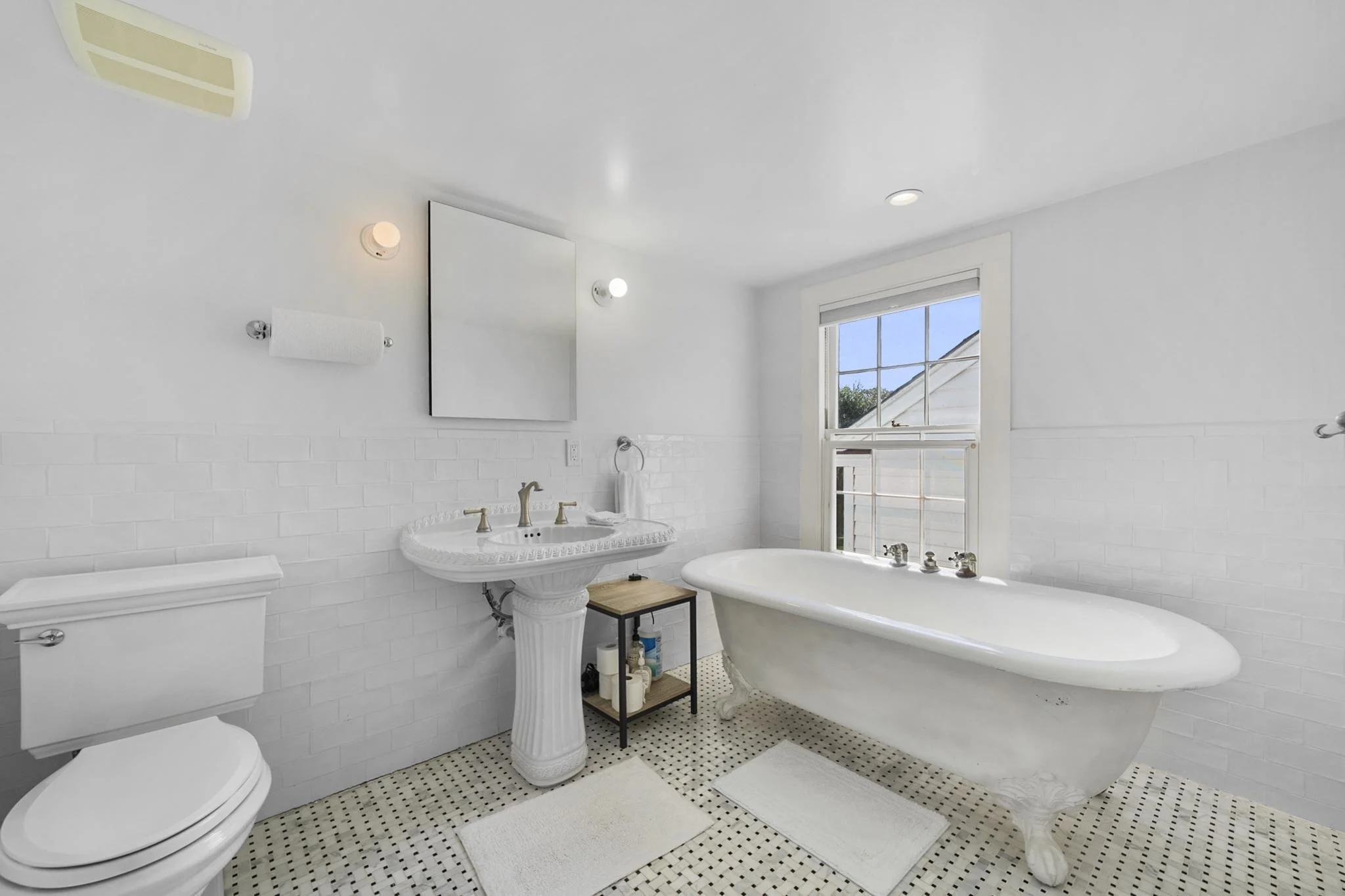 A bright white bathroom featuring a clawfoot bathtub near a window with a view of the outside, a white sink with ornate details, a white toilet, and a small shelf with toiletries. The wall has white subway tiles, and the floor has black and white mos