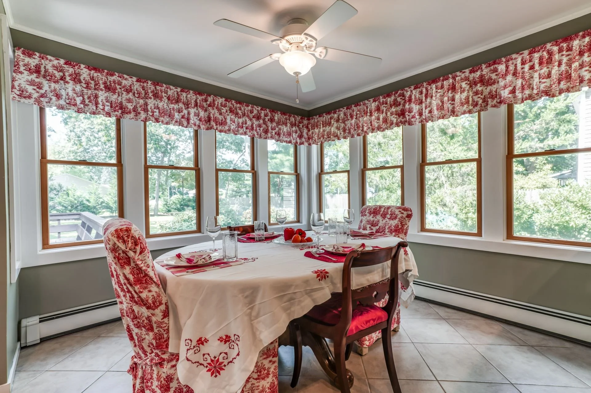 Sunlit dining room with large windows, decorated with red and white toile fabric window valances and chair covers. A round dining table covered with a white tablecloth and red embroidered table runner, set with glassware, plates, napkins, and a cente