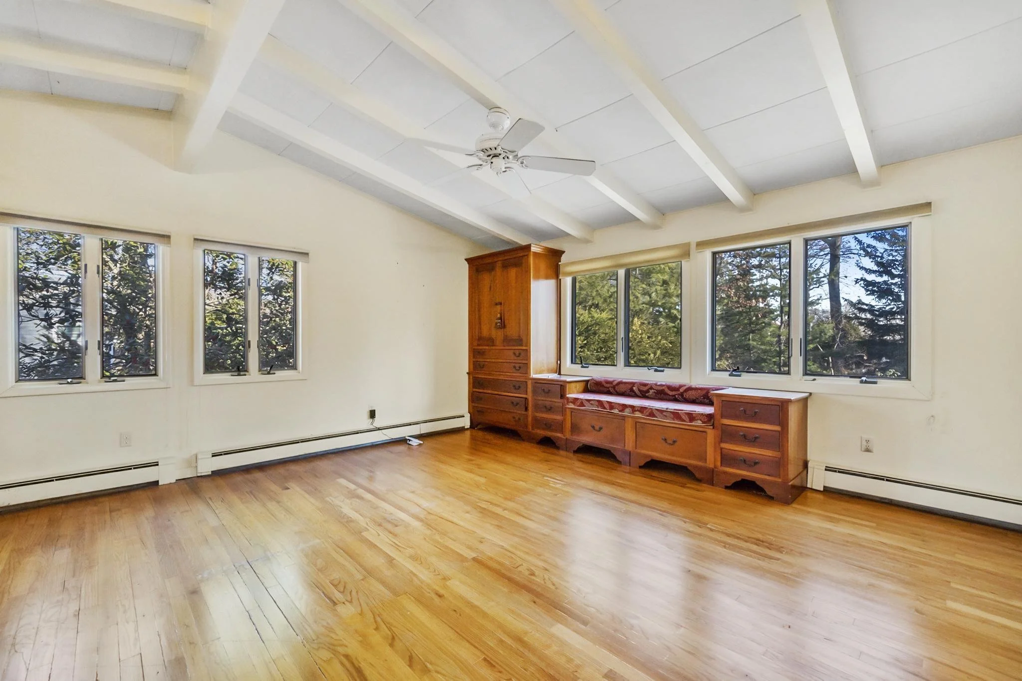Room with wooden floor, cream walls, multiple windows with trees outside, part of ceiling fan visible, built-in wooden seating with drawers.