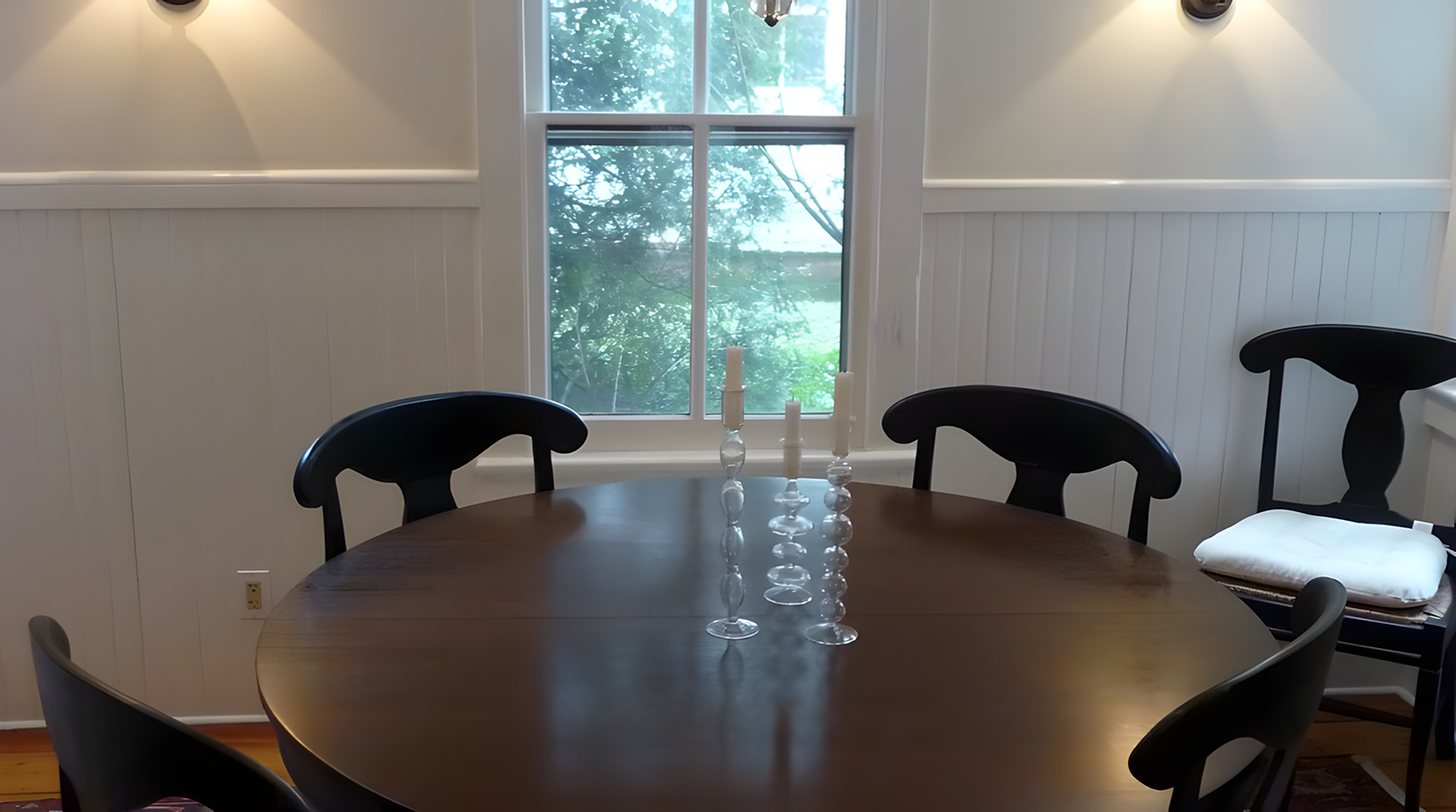 A round wooden dining table with five black chairs, three candles in glass holders, and a window with a view of trees in the background.