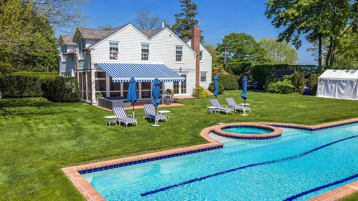 Backyard with swimming pool, hot tub, green lawn, white house, blue and white striped awning, blue lounge chairs, patio furniture, trees, shrubs, and a white tent on a sunny day.