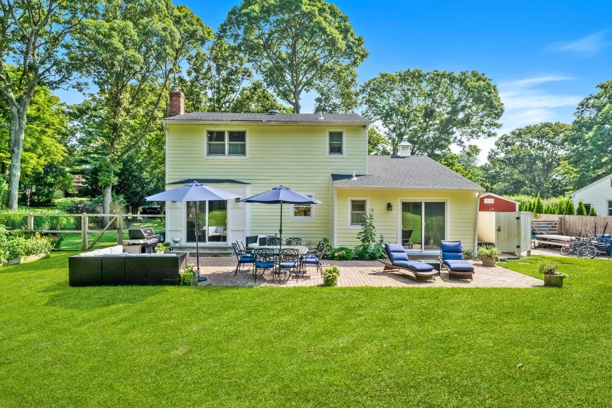 A backyard scene featuring a two-story yellow house with white trim, surrounded by green trees and a well-maintained lawn. The backyard has a patio with outdoor furniture, including tables, chairs, sun loungers, and umbrellas. There is a wooden fence