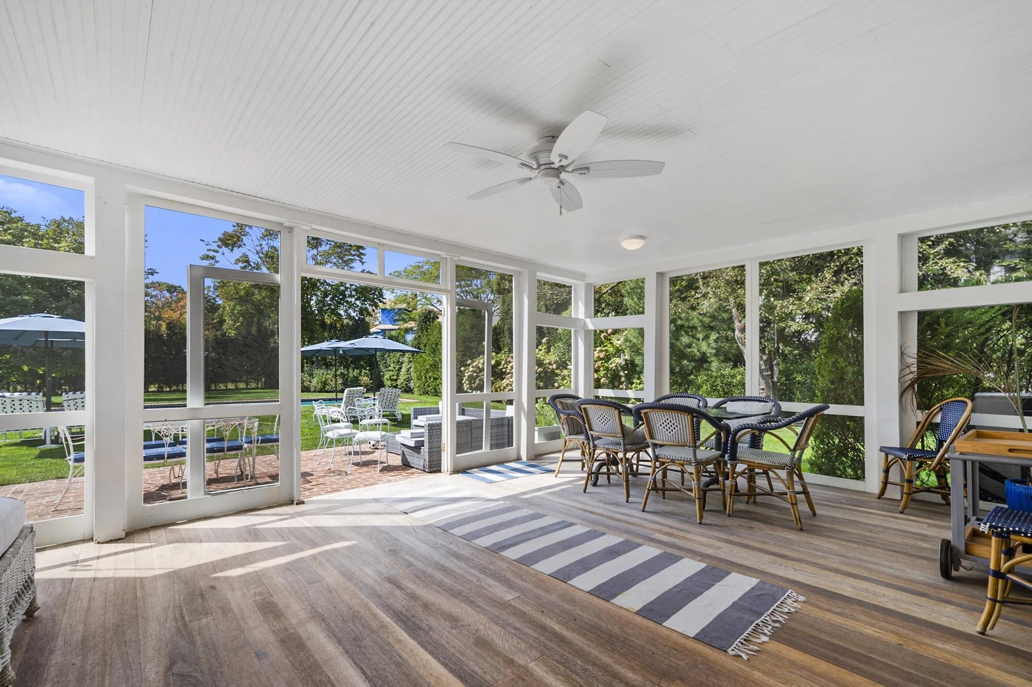 A screened-in porch with wooden flooring, wicker and metal chairs arranged around a table, a striped rug, and views of a backyard with lawn, trees, patio furniture, umbrellas, and a flower bed.