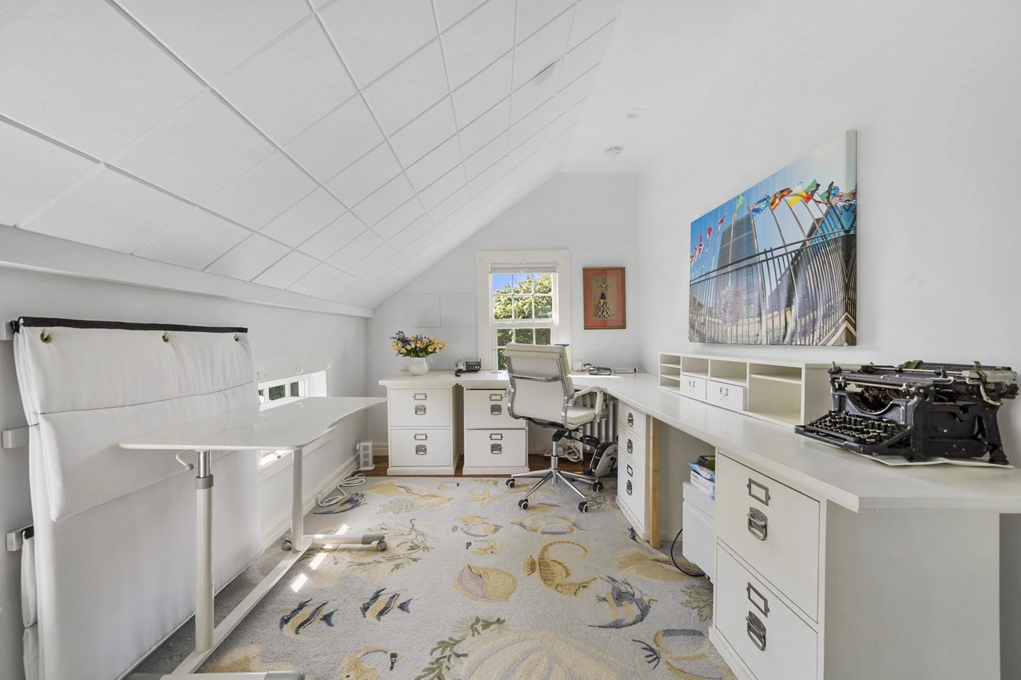 A home office with white walls and furniture, a window with natural light, a desk with a computer, a chair, a telephone, a flower vase, a large colorful carnival photo on the wall, and a vintage typewriter on a side table, with a patterned rug on the