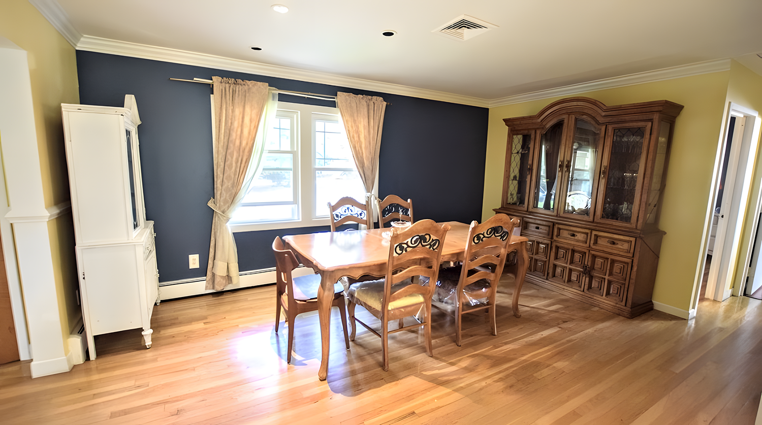 Dining room with a wooden table and six chairs, yellow and dark blue walls, a wooden china cabinet, window with beige curtains, hardwood floor, and ceiling with recessed lighting.