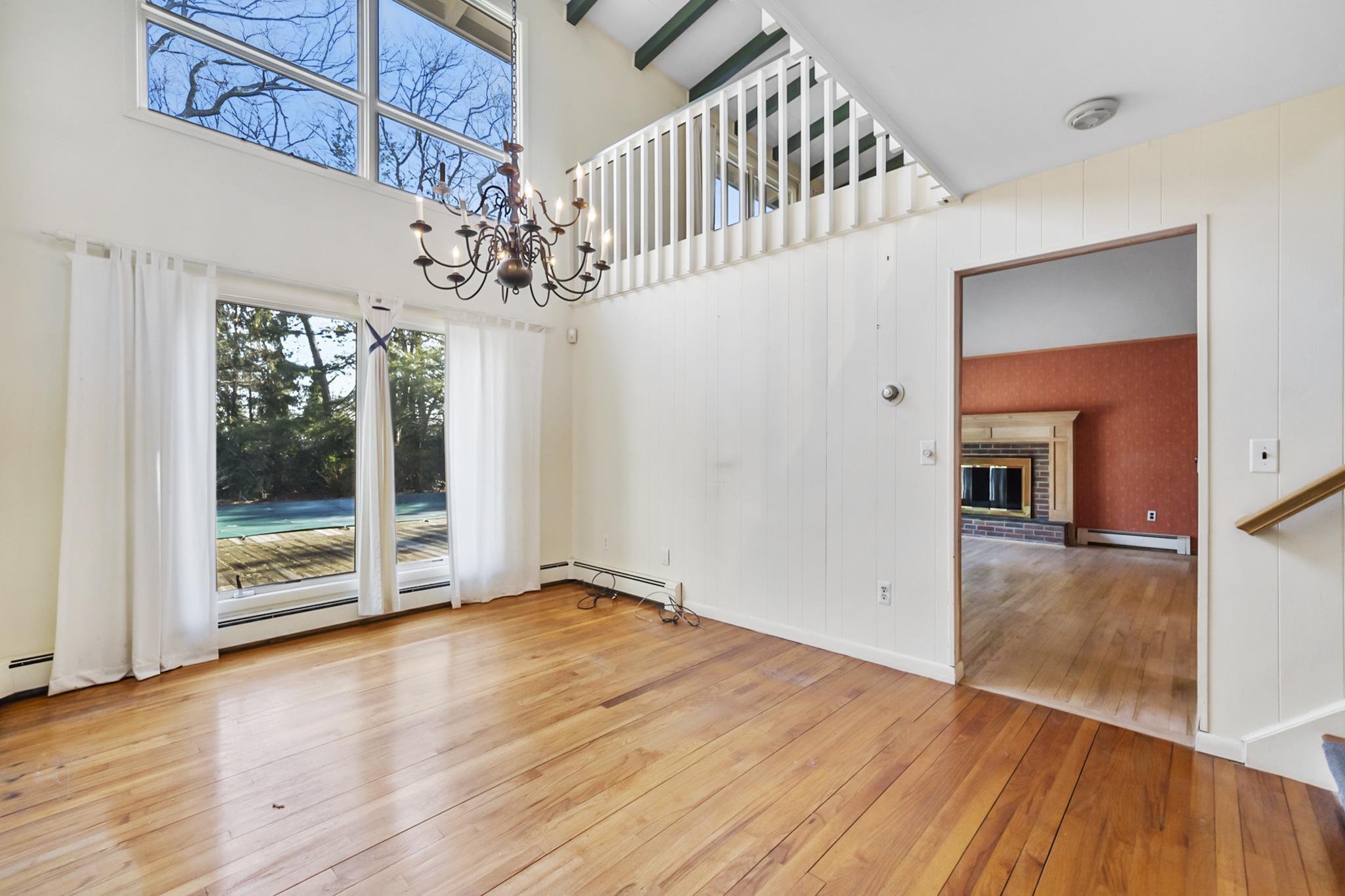 Living room with hardwood flooring, large windows with white curtains, and a chandelier hanging from the ceiling. An opening leads to another room with a red wall and a fireplace.