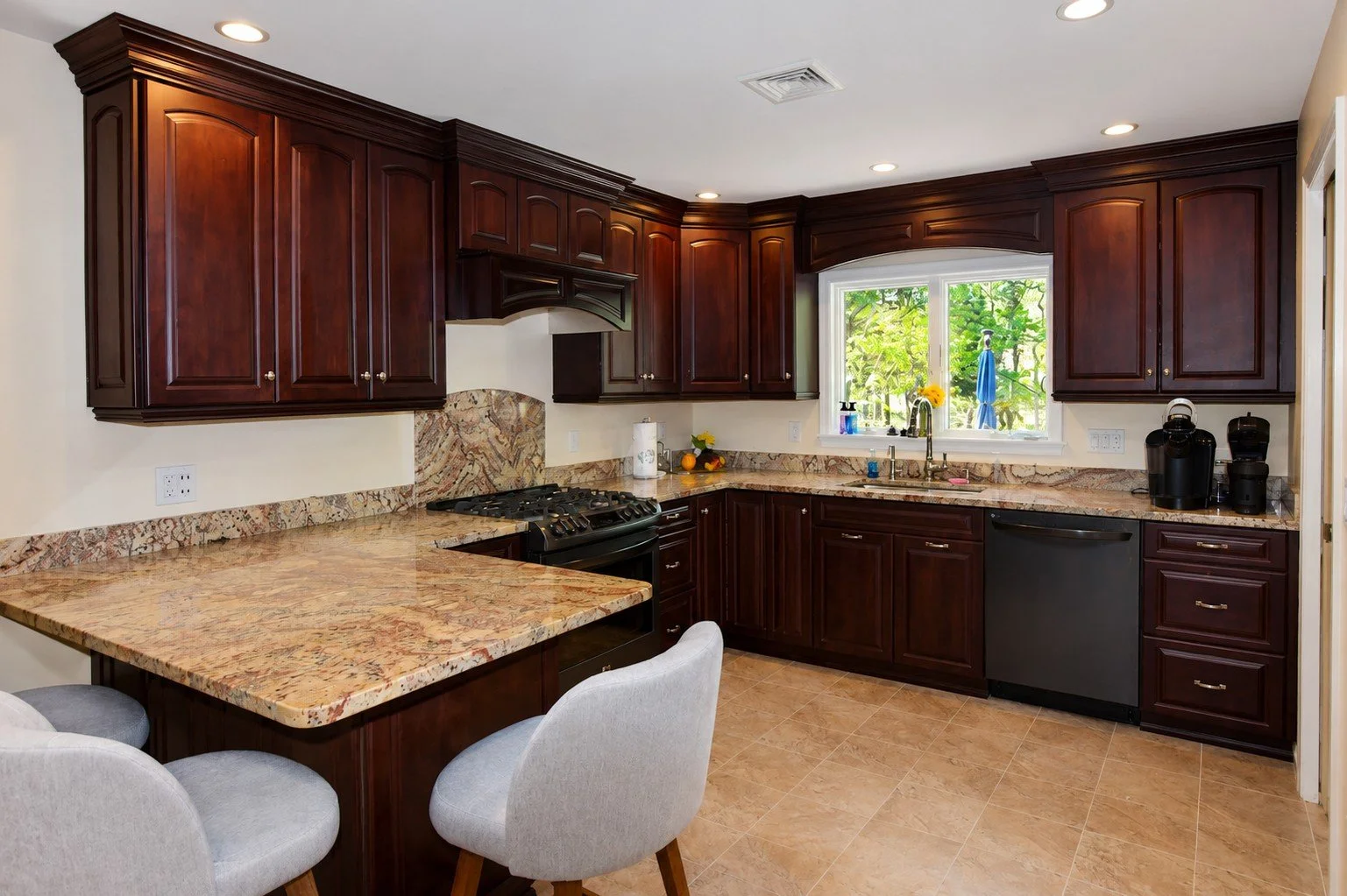 A kitchen with dark wood cabinets, marble countertops, a window with greenery outside, a stove, coffee maker, and dining area with light-colored chairs.