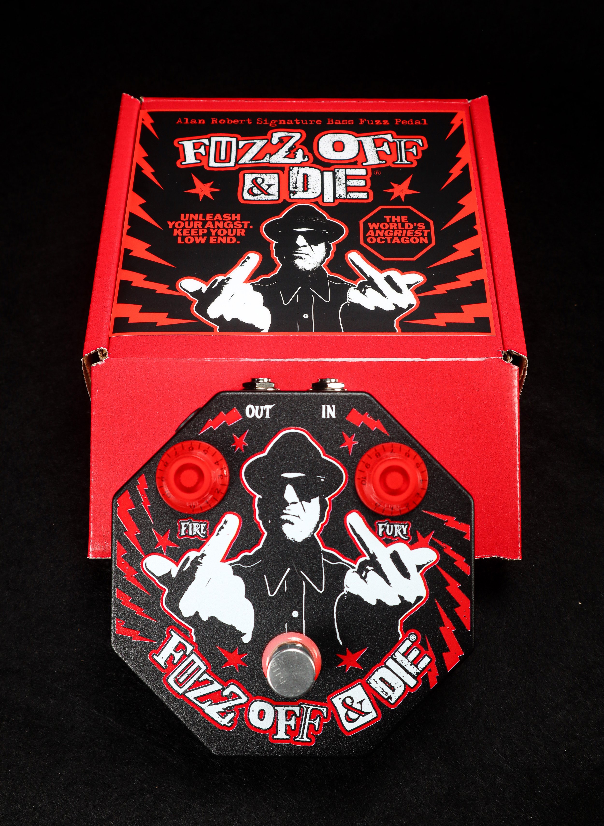 A black, red, and white guitar effects pedal named FUZZ OFF & DIE with an illustration of a man in black sunglasses and hat, showing middle fingers. The pedal has two red knobs labeled 'FIRE' and 'FURY', and an input and output jack labeled 'IN' and 'OUT'. It is placed in front of a red and black box with similar graphics and text.