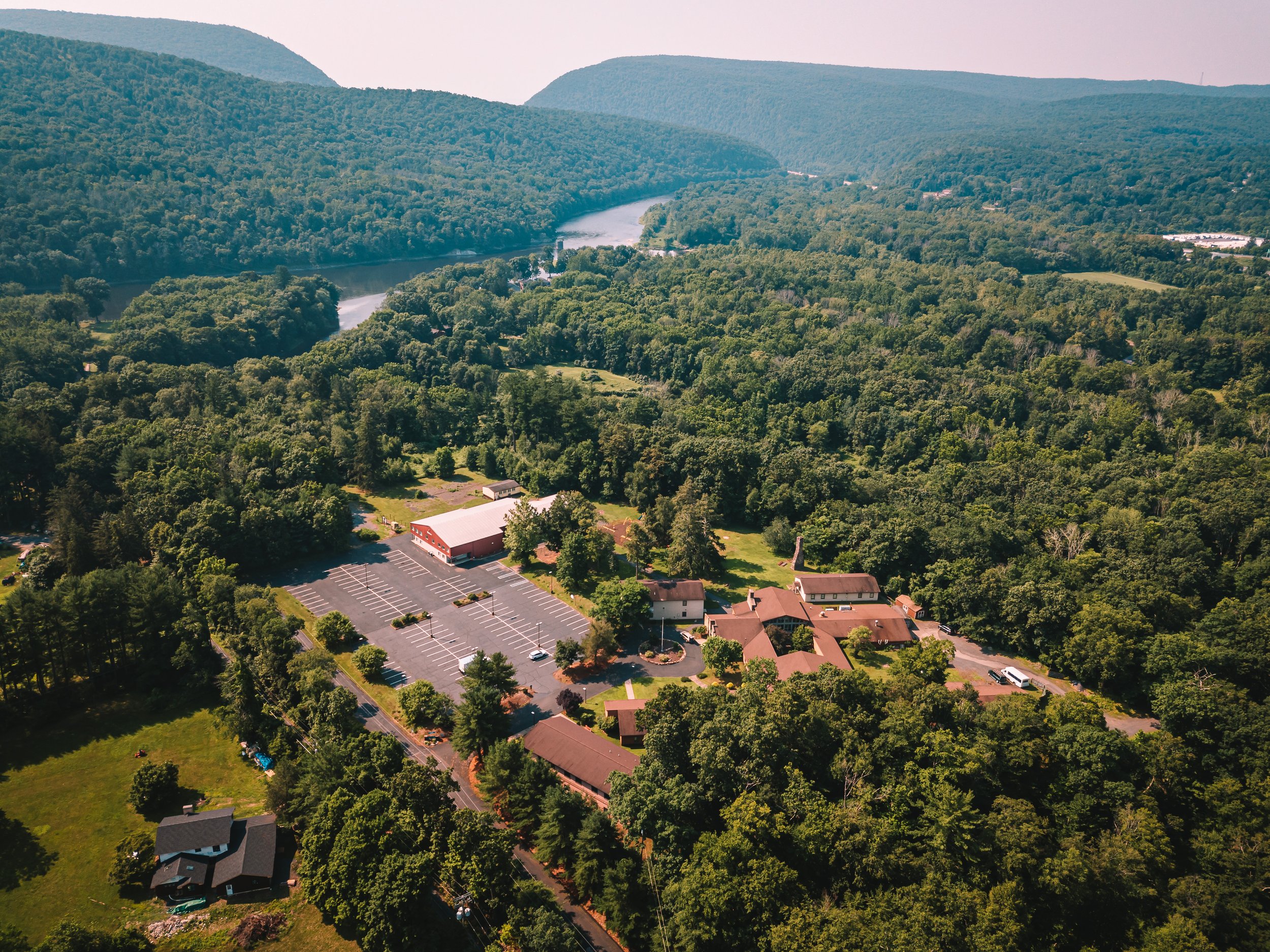 Aerial view of a rural area with a large parking lot surrounded by trees, a few buildings, and a river winding through a green landscape with hills in the background.