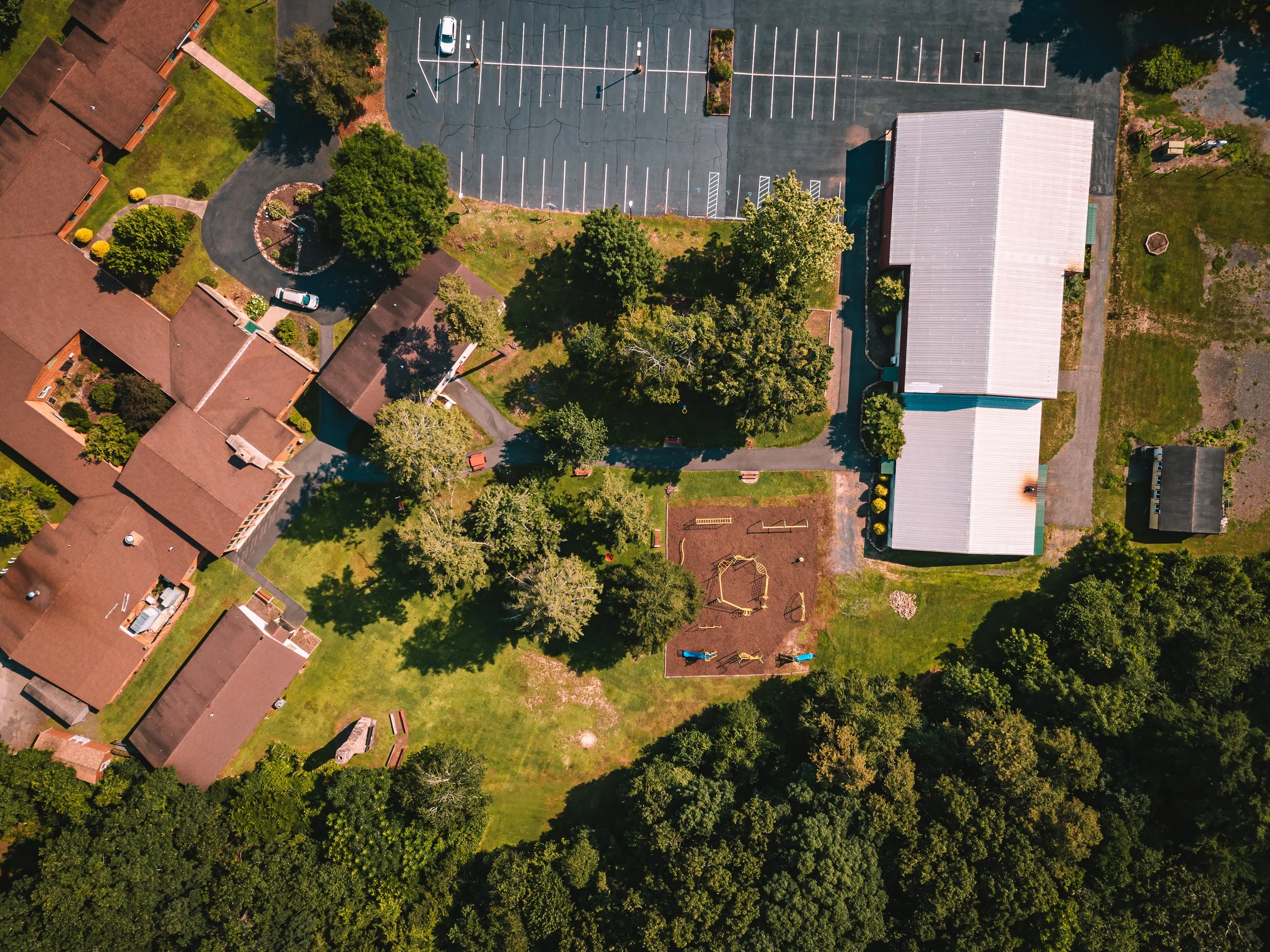 Aerial view of a residential area with several houses, a playground, trees, a parking lot, and a large building with a metal roof.