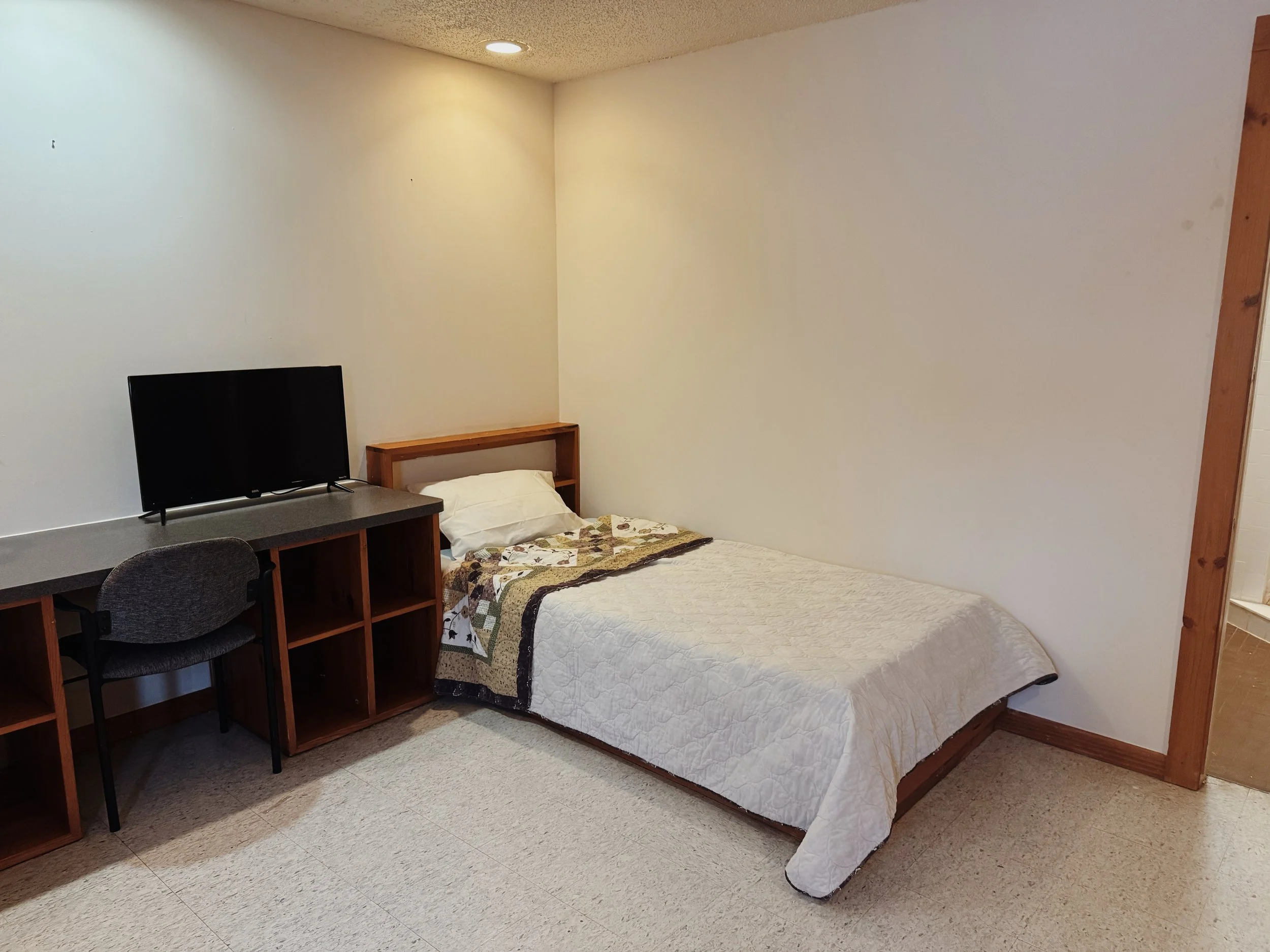 A small room with a single bed, white quilt, and pillow. Next to the bed is a wooden shelf headboard. A flat-screen TV sits on a dark desk with open cubbies. A gray cushioned chair is in front of the desk. The room has beige carpet, plain white walls, and a doorway leading to a bathroom.