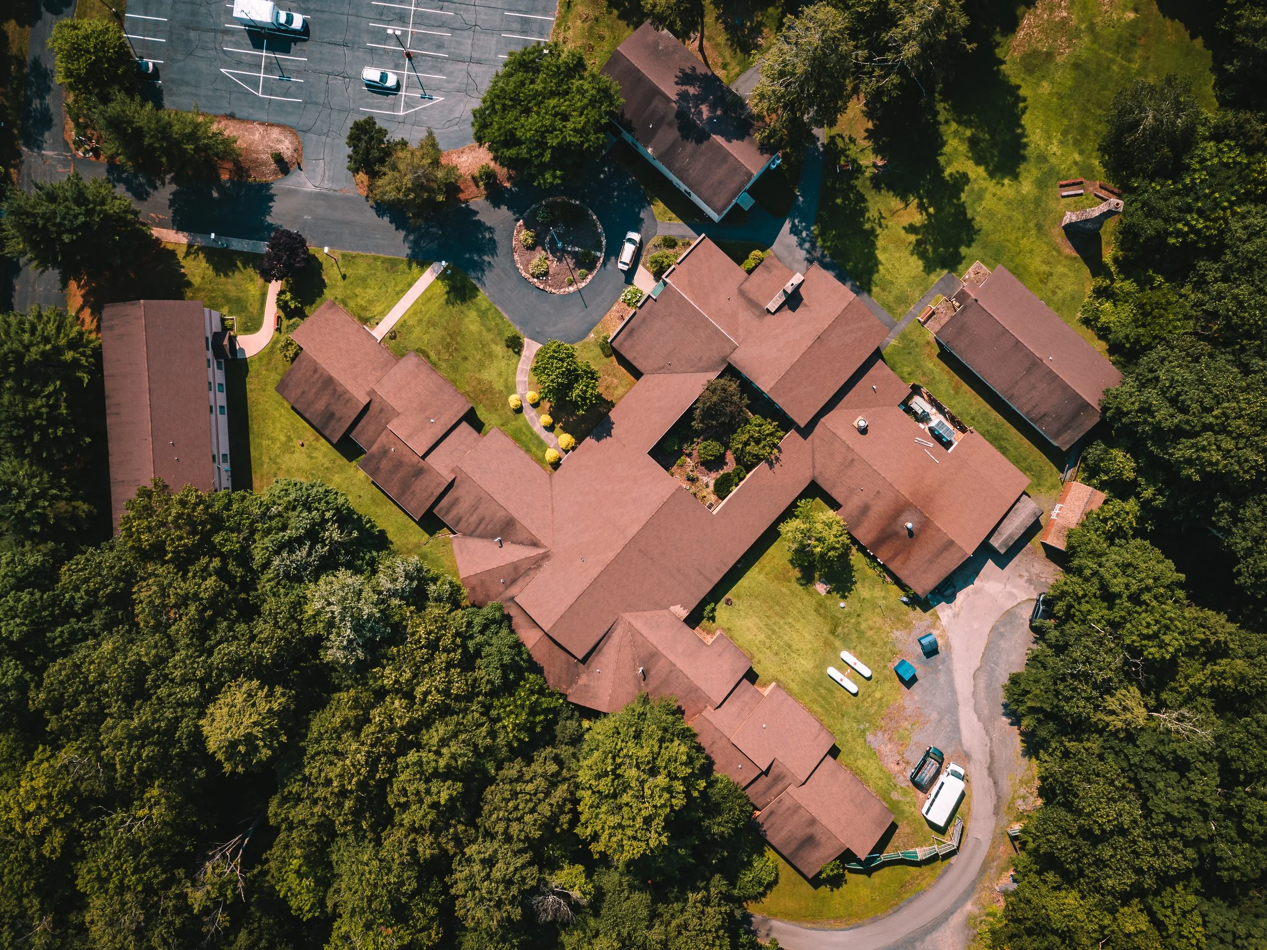 An aerial view of a residential area with several houses, green trees, a parking lot, and a small tennis court surrounded by greenery.