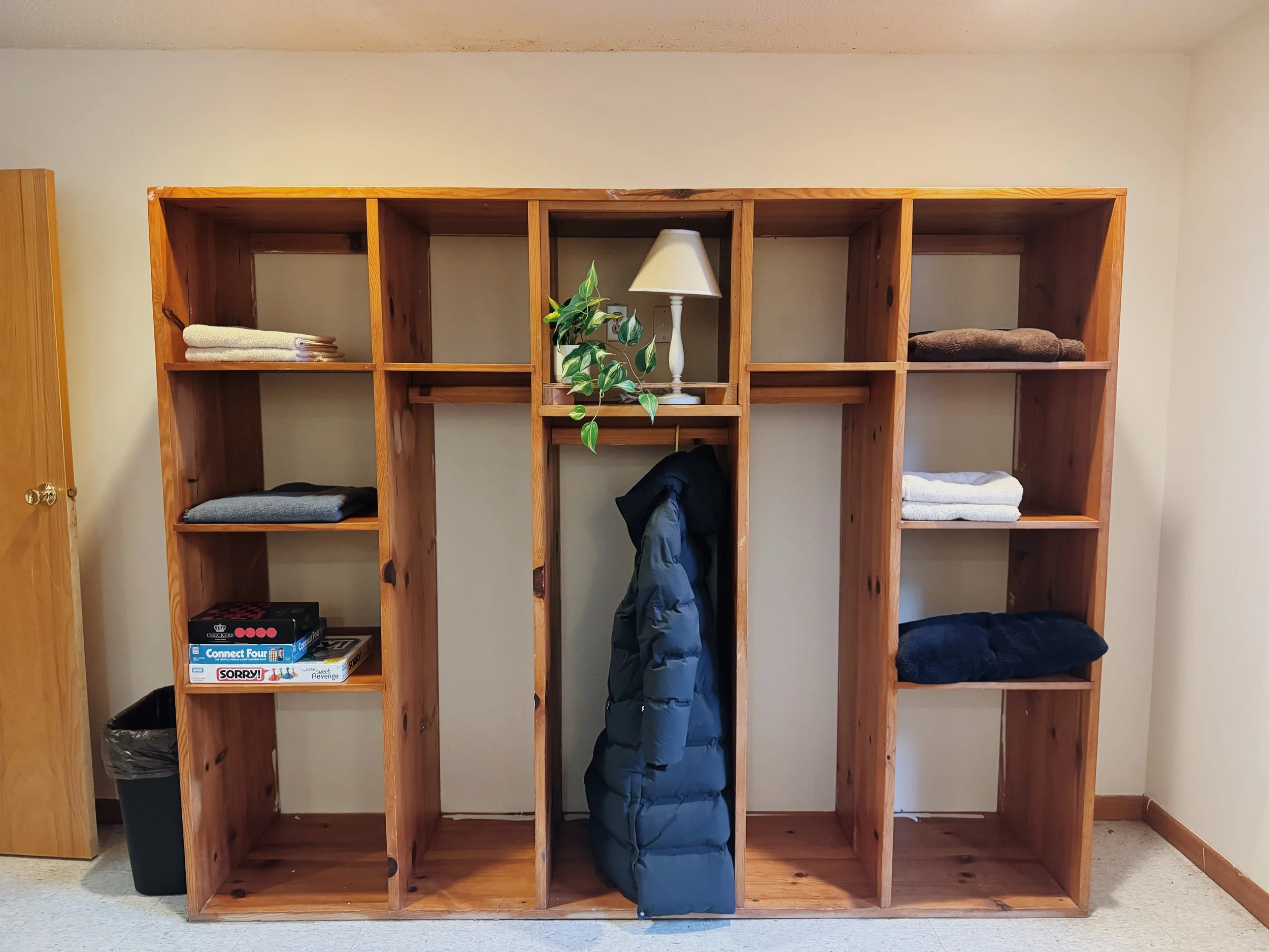 Large wooden shelf with various folded towels, a black coat hanging, a dresser, a potted plant, a white table lamp, and a black trash can.