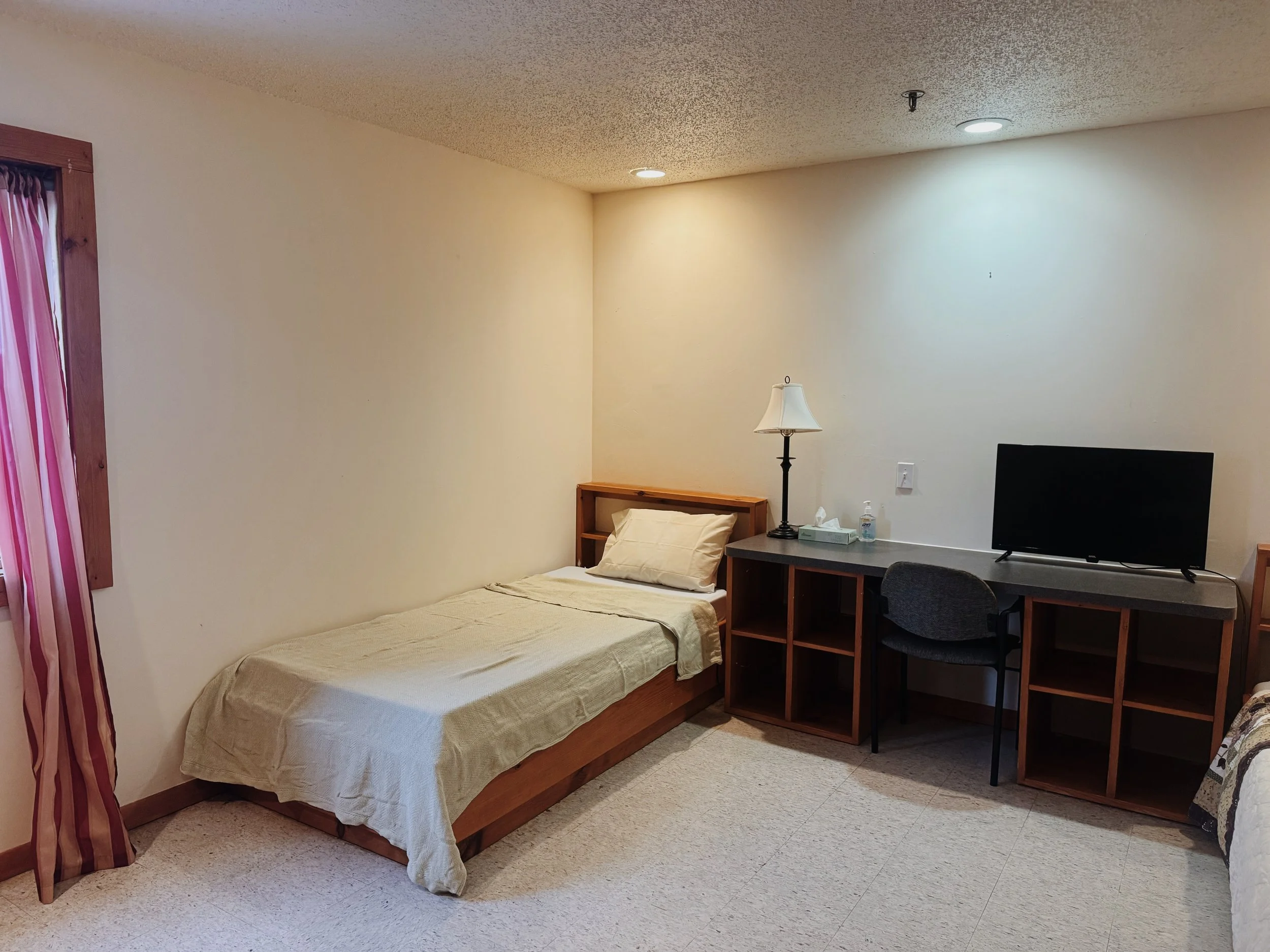 Room with a single bed, a desk with a TV, a lamp, and some toiletries, beige walls, carpeted floor, and a window with striped curtains.
