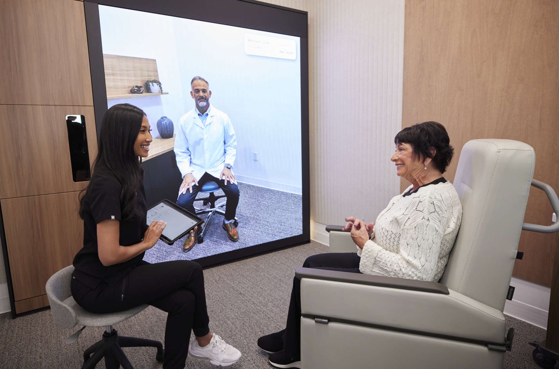 A woman in a black outfit sitting on an office chair, holding a tablet, talking to an elderly woman sitting in a chair, in a medical or consultation room. A large screen behind them displays a man in a white coat, sitting on a stool, in a virtual consultation.