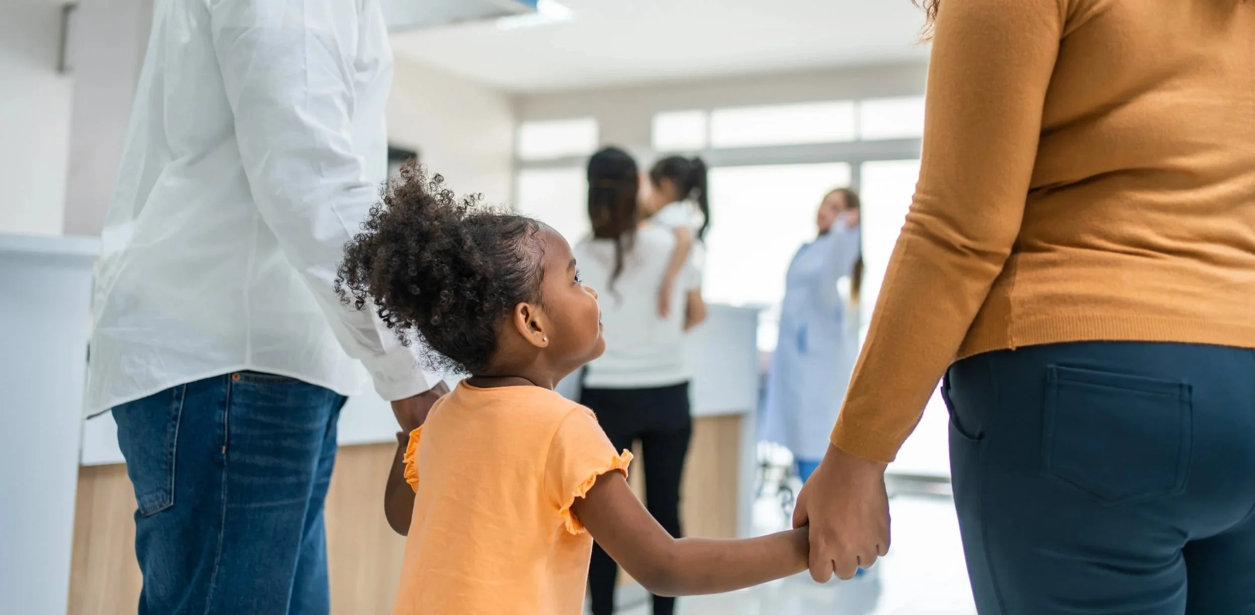 A young girl is holding hands with a woman in a medical facility, with other children and healthcare workers in the background.