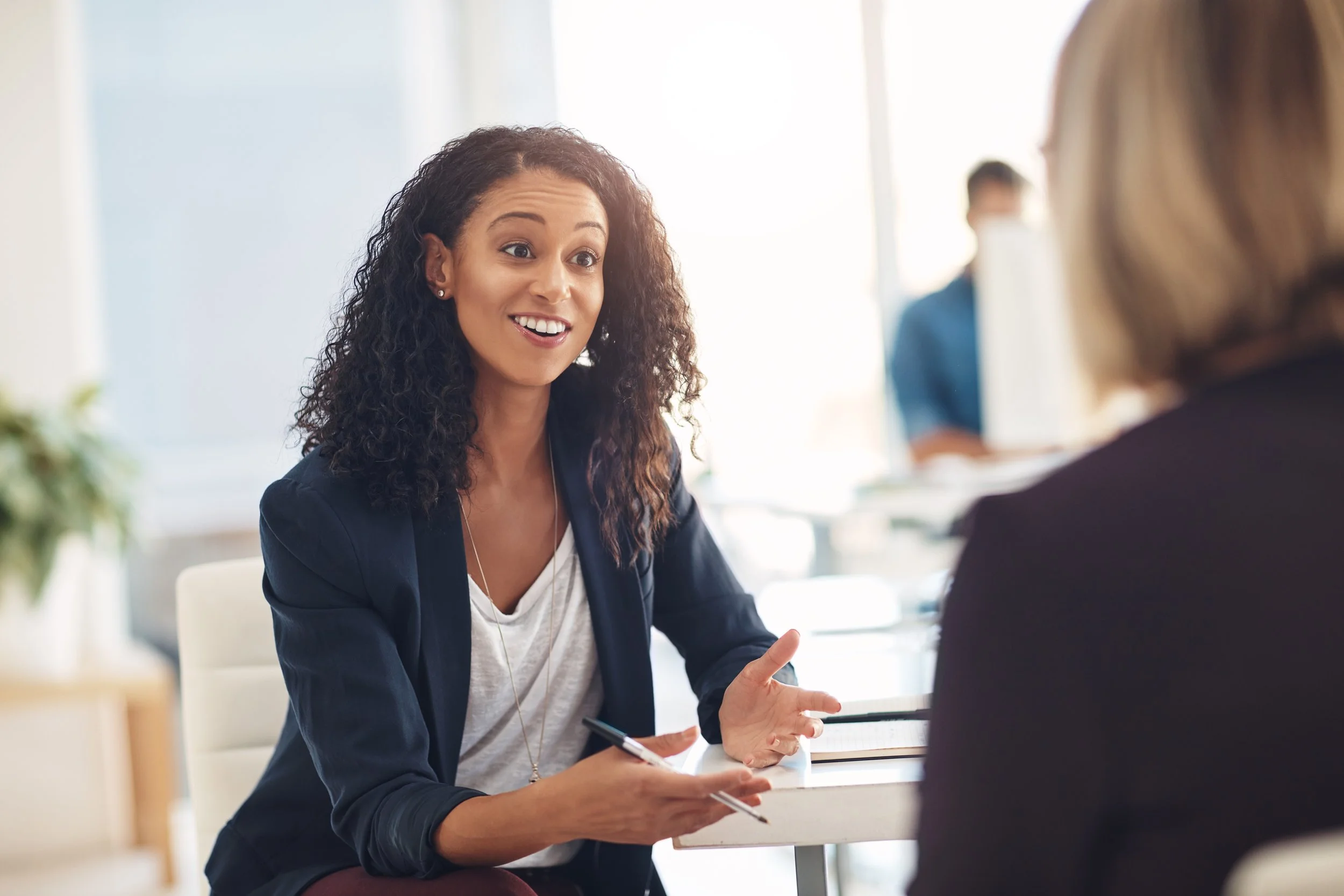 A woman with curly hair, wearing a navy blazer and a white top, engaged in a conversation with another woman in an office setting.