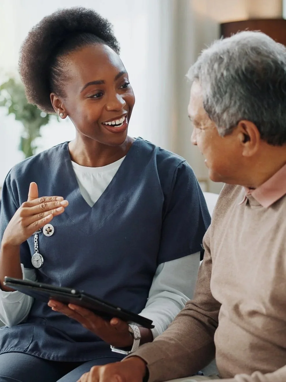 A young female healthcare professional in a blue uniform, holding a tablet, engaged in a conversation with an elderly man in a beige shirt, both smiling and appearing to have a friendly discussion inside a well-lit room.