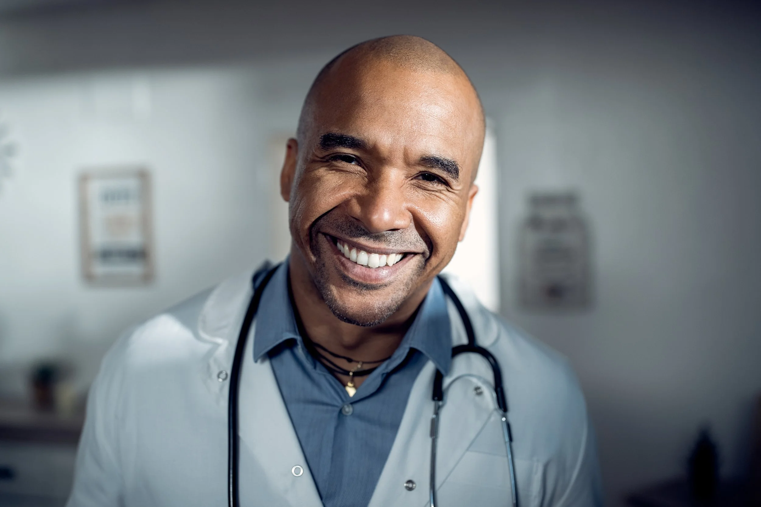 A smiling male doctor with a stethoscope around his neck in a medical setting.