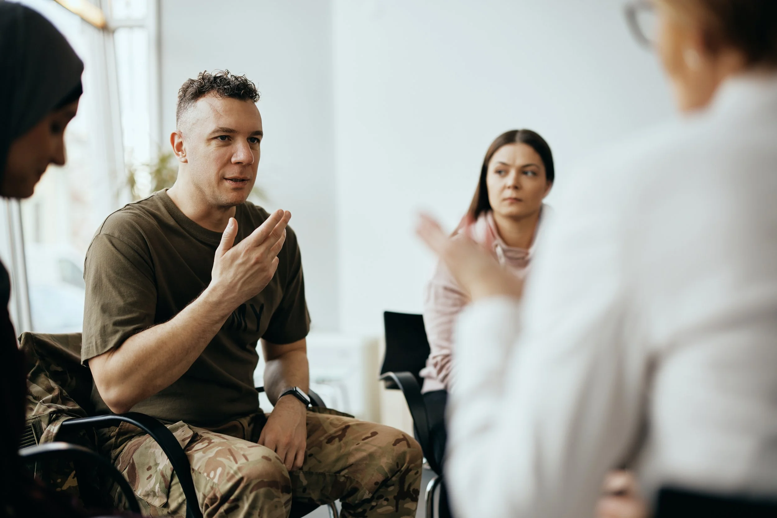 A man in military camouflage pants and a brown t-shirt is speaking and gesturing with his hand. He is in a group therapy session with a woman in a pink hoodie and a woman in a white coat, possibly a therapist, in a brightly lit room.