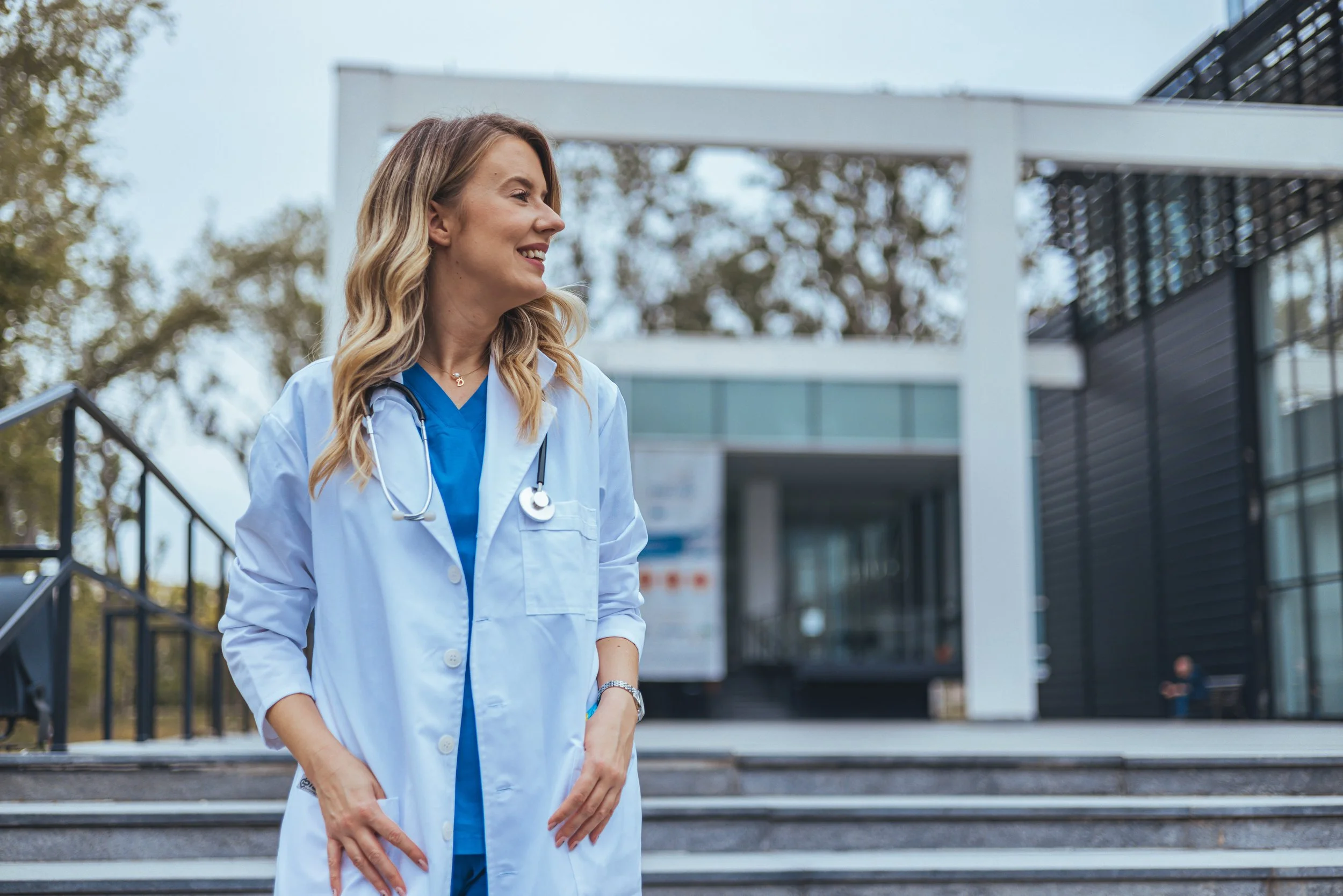 A female healthcare professional wearing a white coat and blue scrubs with a stethoscope around her neck, standing outdoors on steps in front of a modern building, smiling and looking to the side.