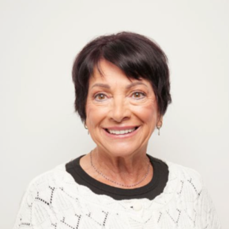 Smiling older woman with short dark hair wearing a white knit sweater with black accents, standing against a neutral background.