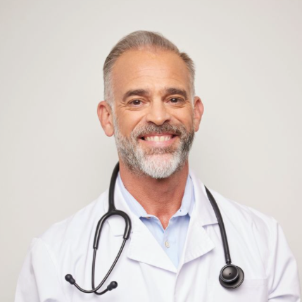 A smiling male doctor with gray hair, a beard, and a stethoscope around his neck, wearing a white coat against a plain background.