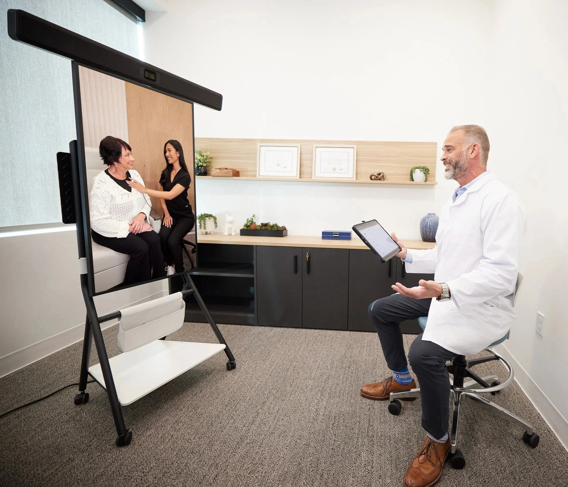 A doctor conducting a virtual consultation with two women, one older and one younger, in a medical office with a computer display showing the women. The doctor is seated, holding a tablet, and smiling.